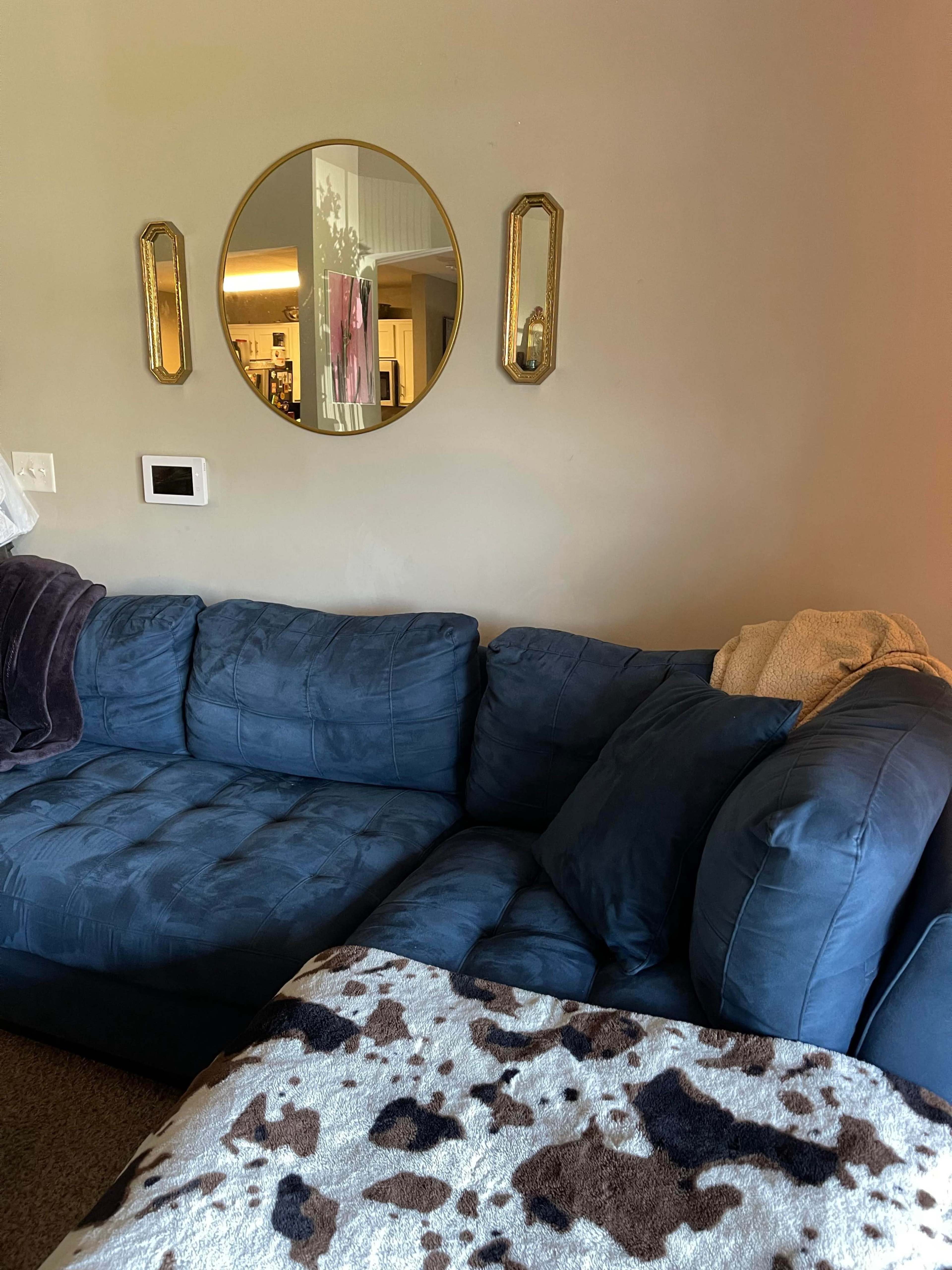 The image shows a blue sectional sofa in a living room with a round mirror and two wall sconces hanging above it.