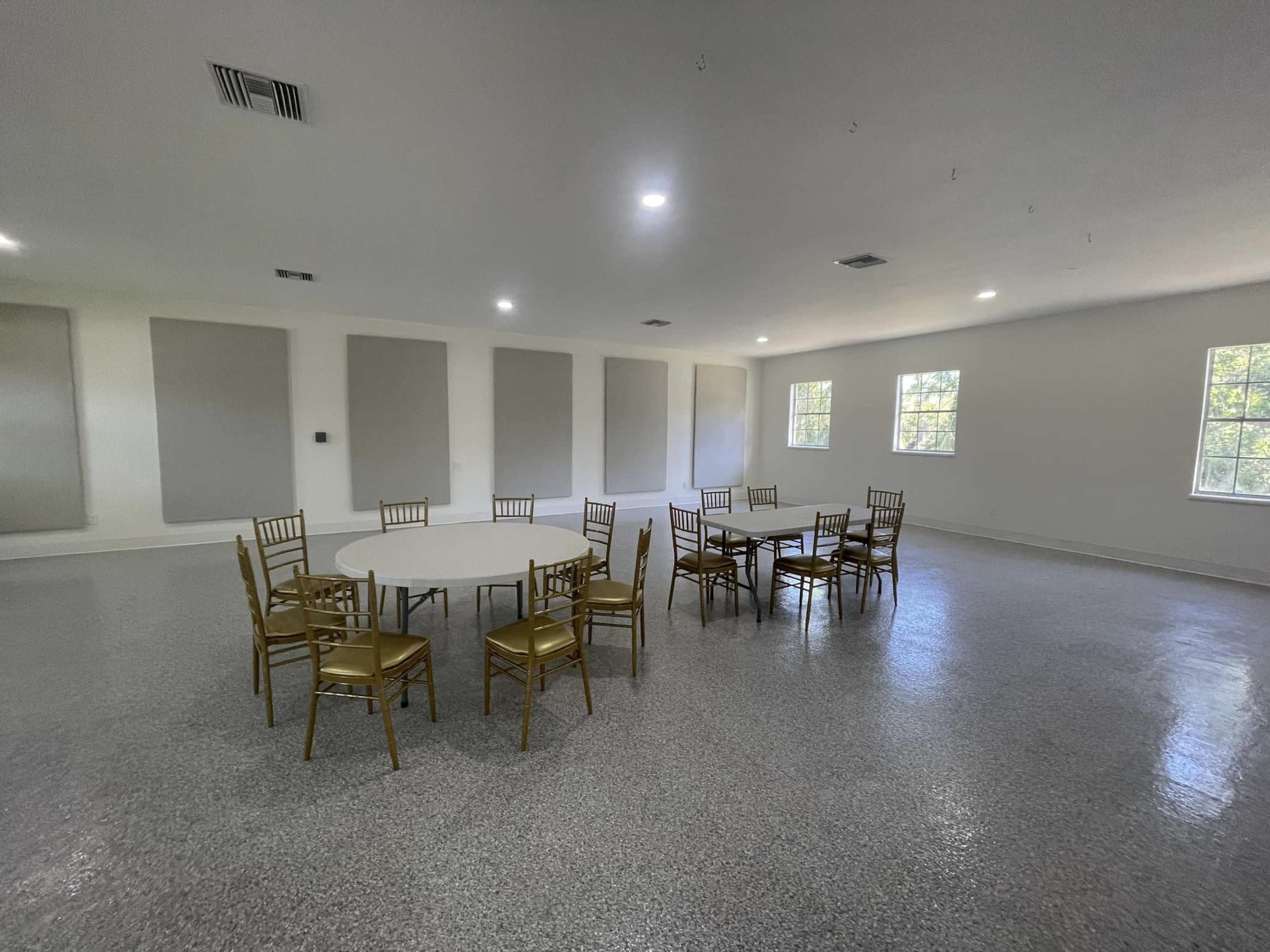 A spacious room features several round tables surrounded by gold chairs, with windows allowing natural light to enter.