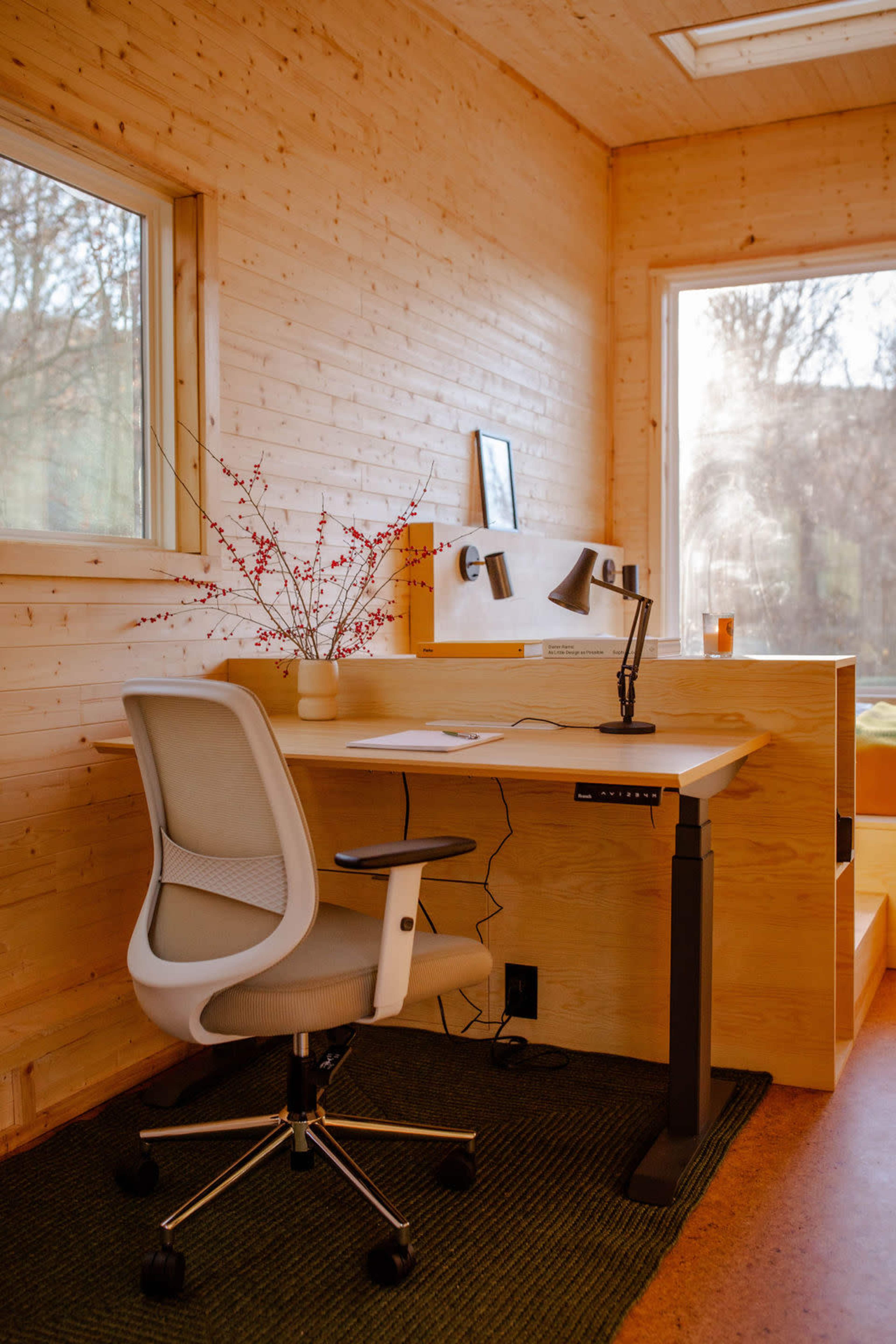 A simple office space with a wooden desk, ergonomic chair, lamp, and a vase with branches, set against a backdrop of large windows.