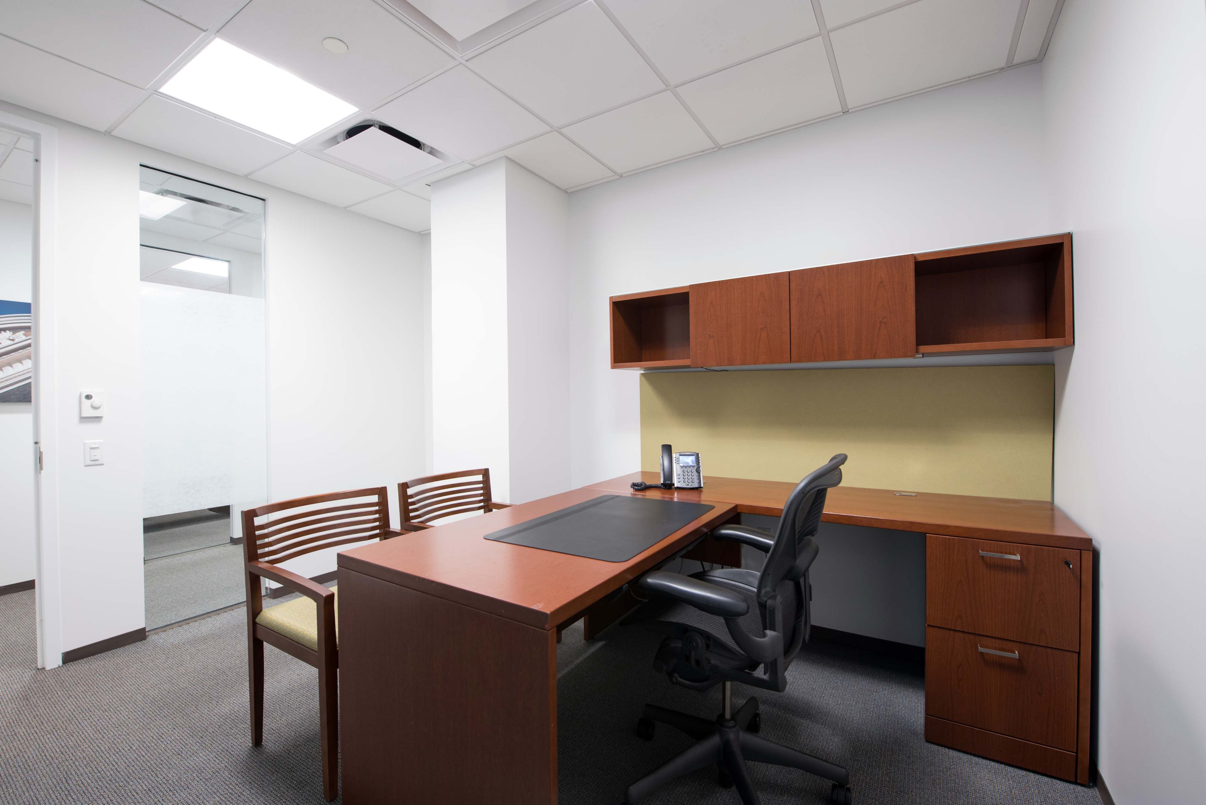 A minimalist office space featuring a wooden desk, a black ergonomic chair, a wall-mounted shelf, and two wooden chairs.