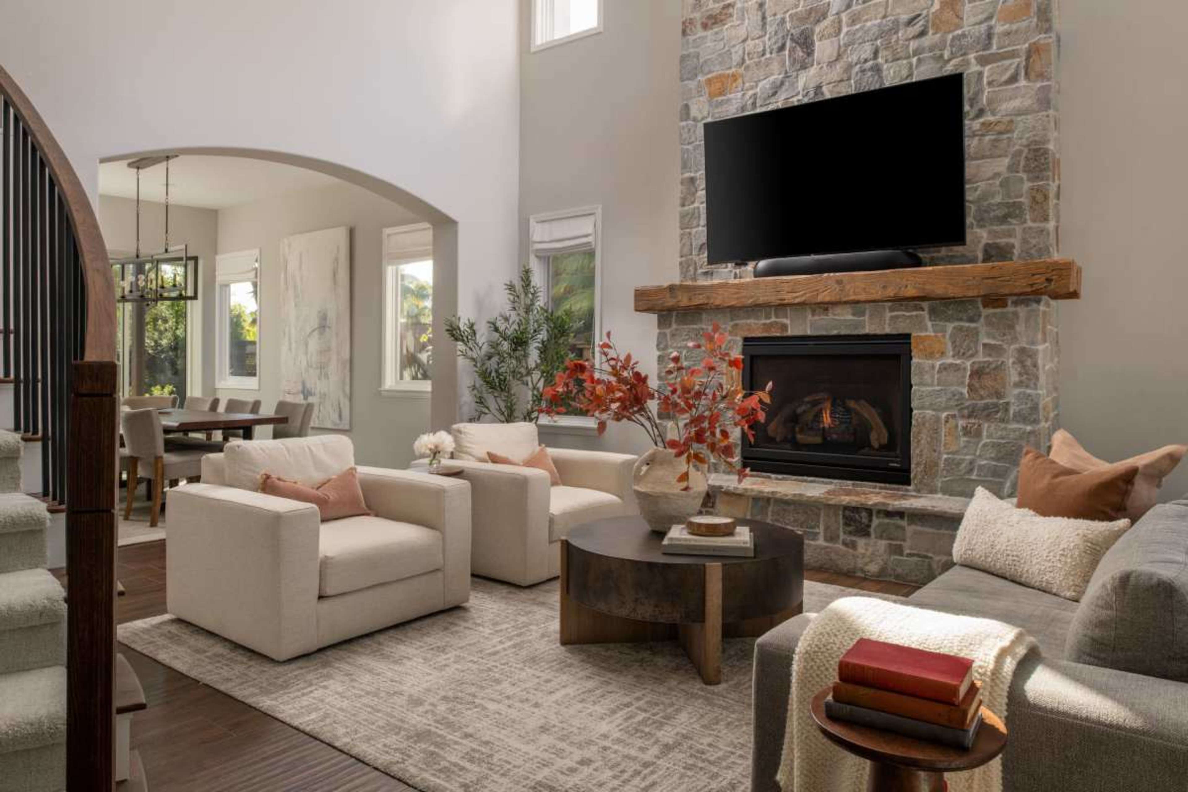 The living room features a stone fireplace with a mounted television above, surrounded by two white armchairs and a coffee table, all set on a textured area rug.