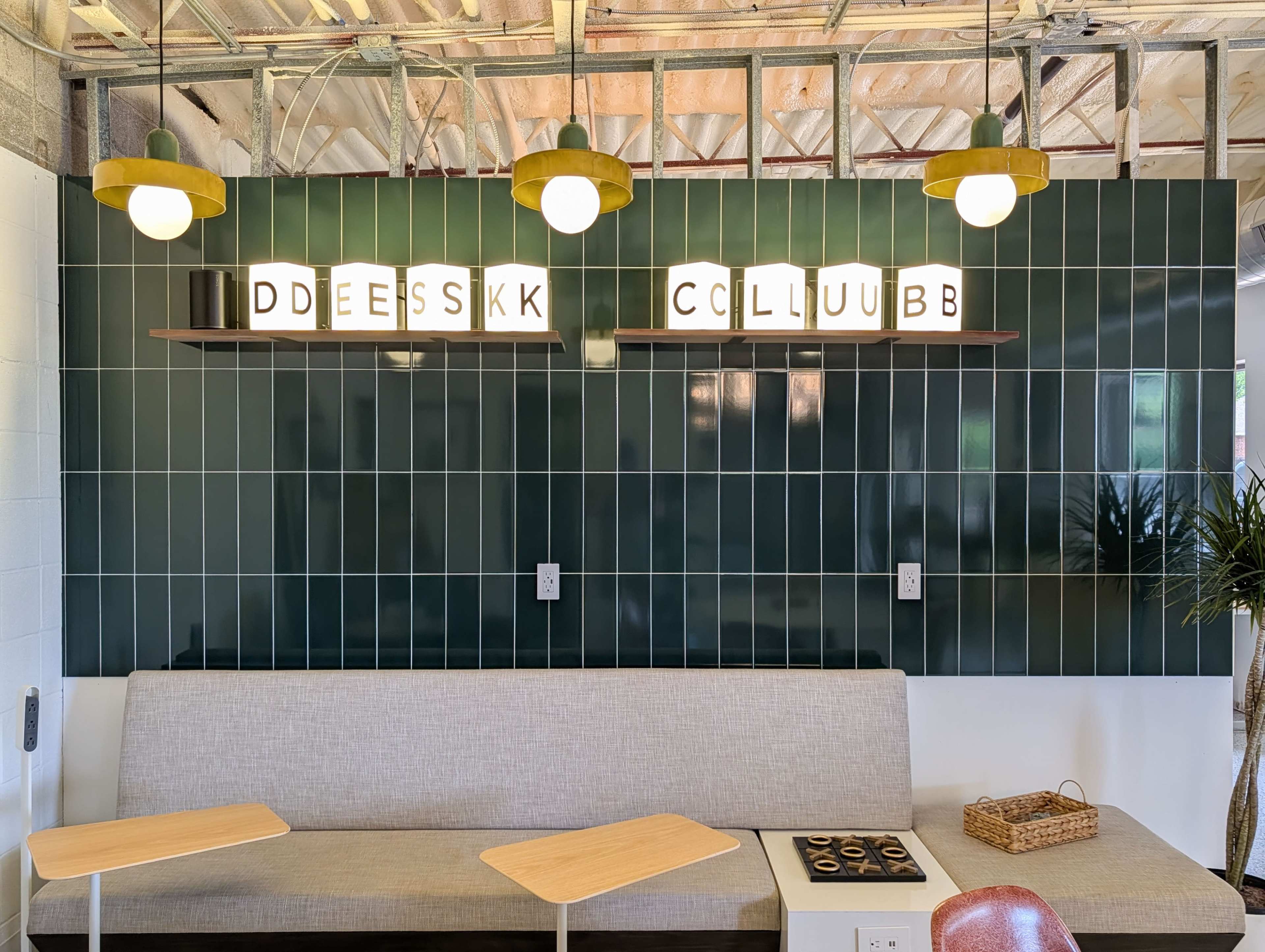 The image shows a modern café interior with a green tiled wall, a long gray bench, and a shelf displaying illuminated letter blocks.
