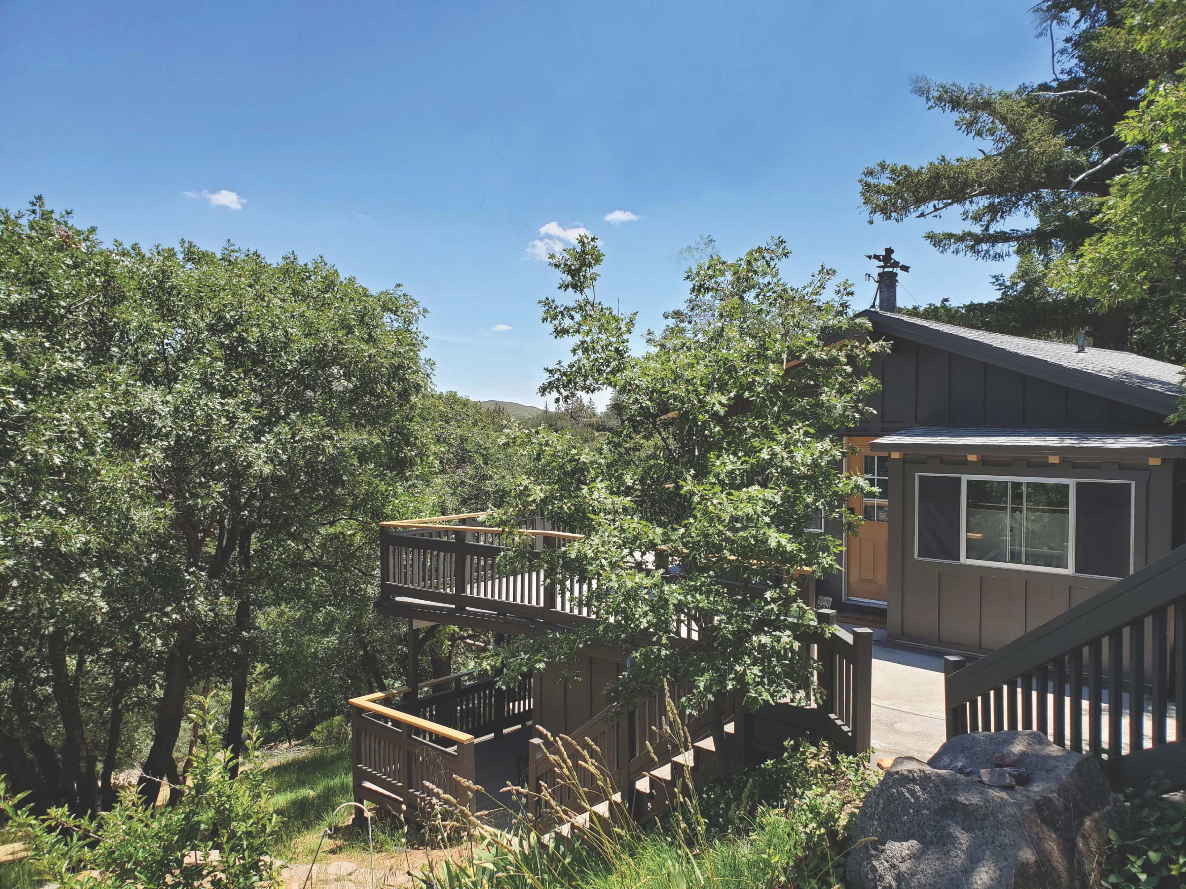 A two-story wooden house is situated among trees on a hillside, featuring a deck that extends from the side of the building.
