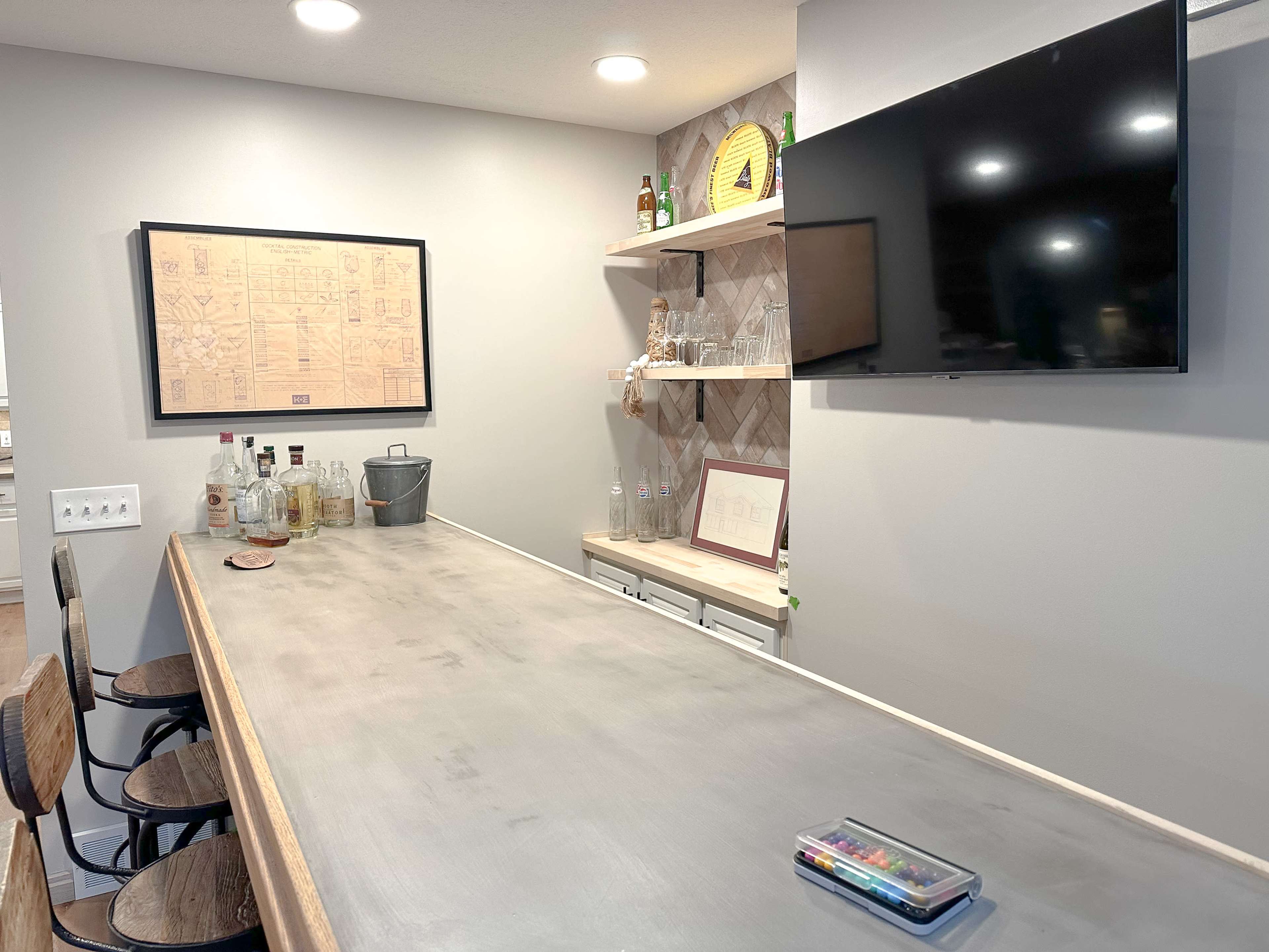 A home bar area with a concrete countertop, shelves displaying bottles, and a television mounted on the wall.