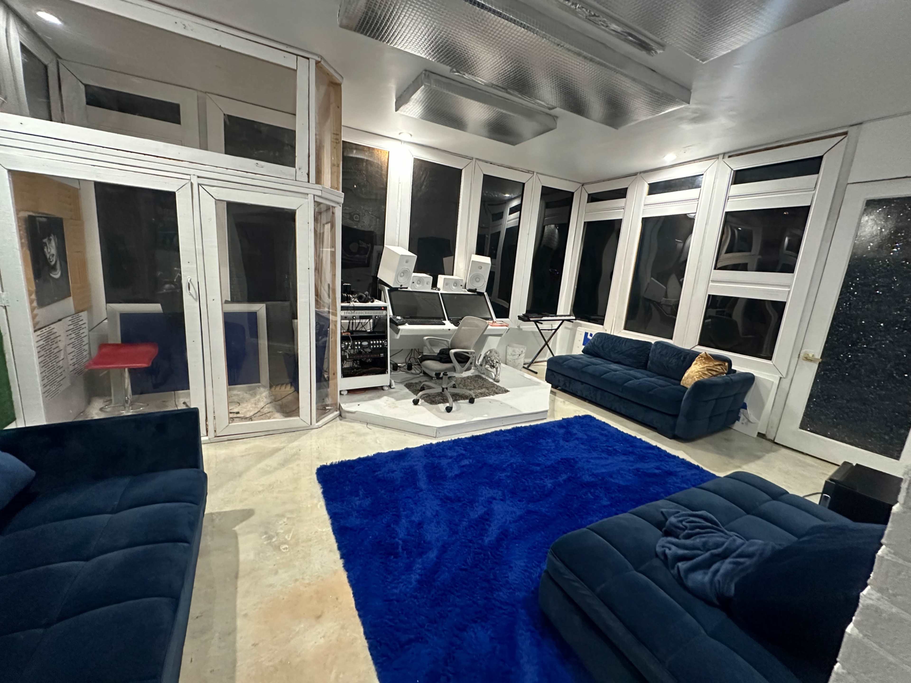 The image shows a modern room featuring large windows, two dark blue sofas, a blue rug, and a workstation with multiple monitors.