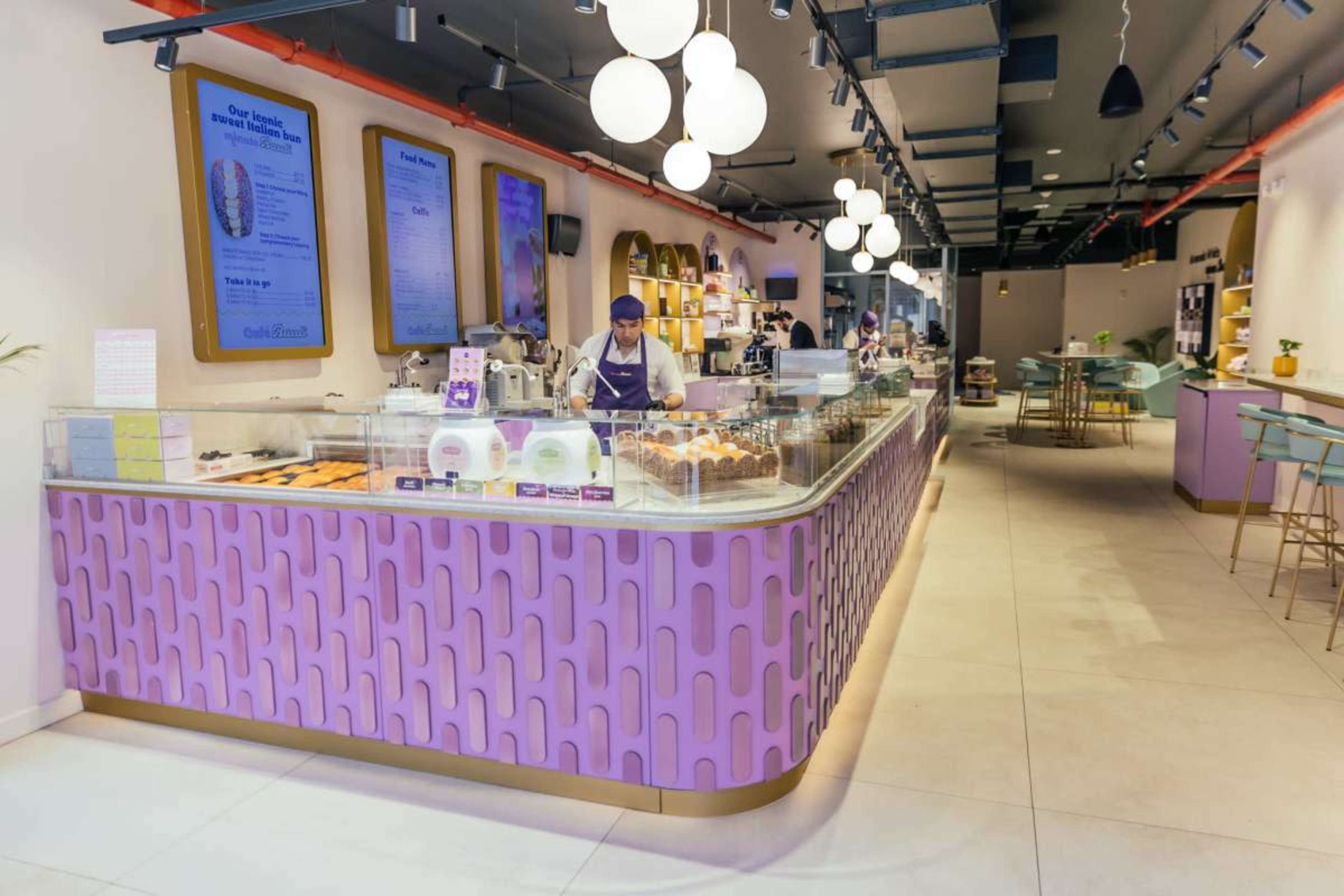 A modern dessert shop features a vibrant purple and gold counter, with a barista preparing items behind glass displays.