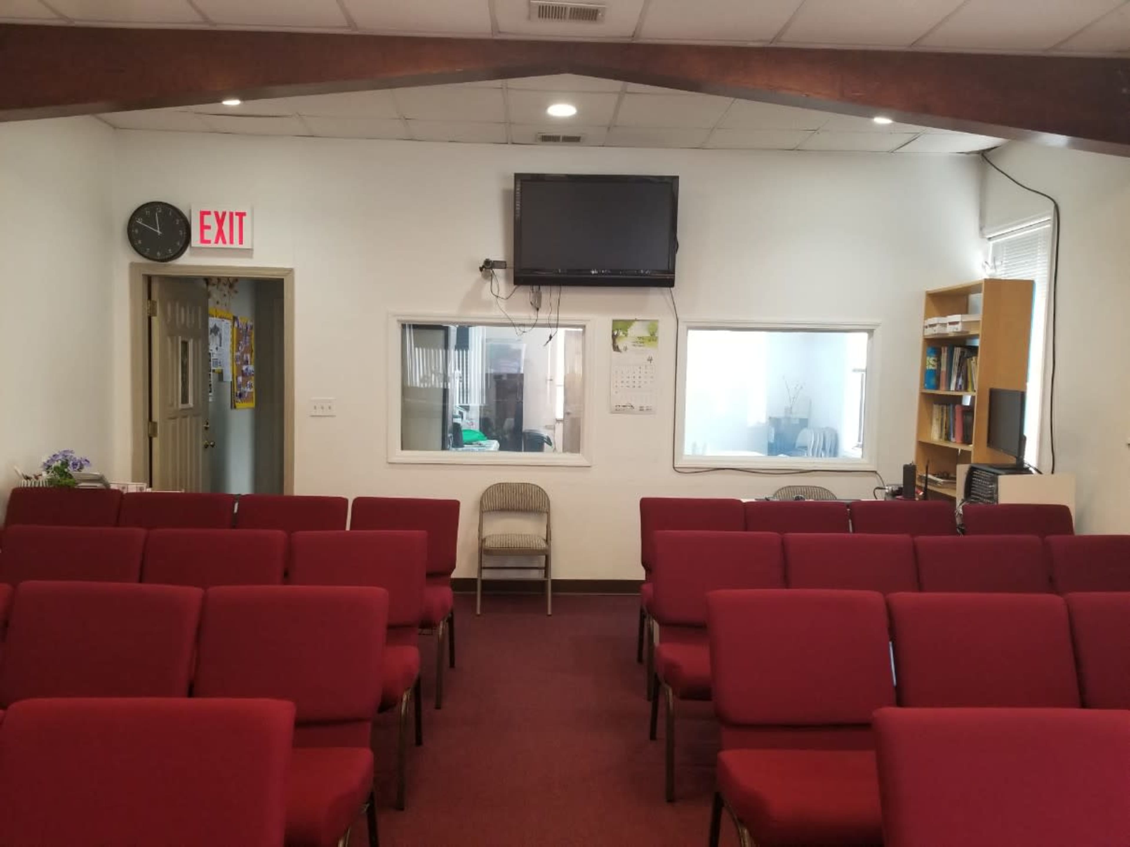 The image shows a room with several rows of red chairs facing a wall, with a television mounted above and two windows on either side of a door.