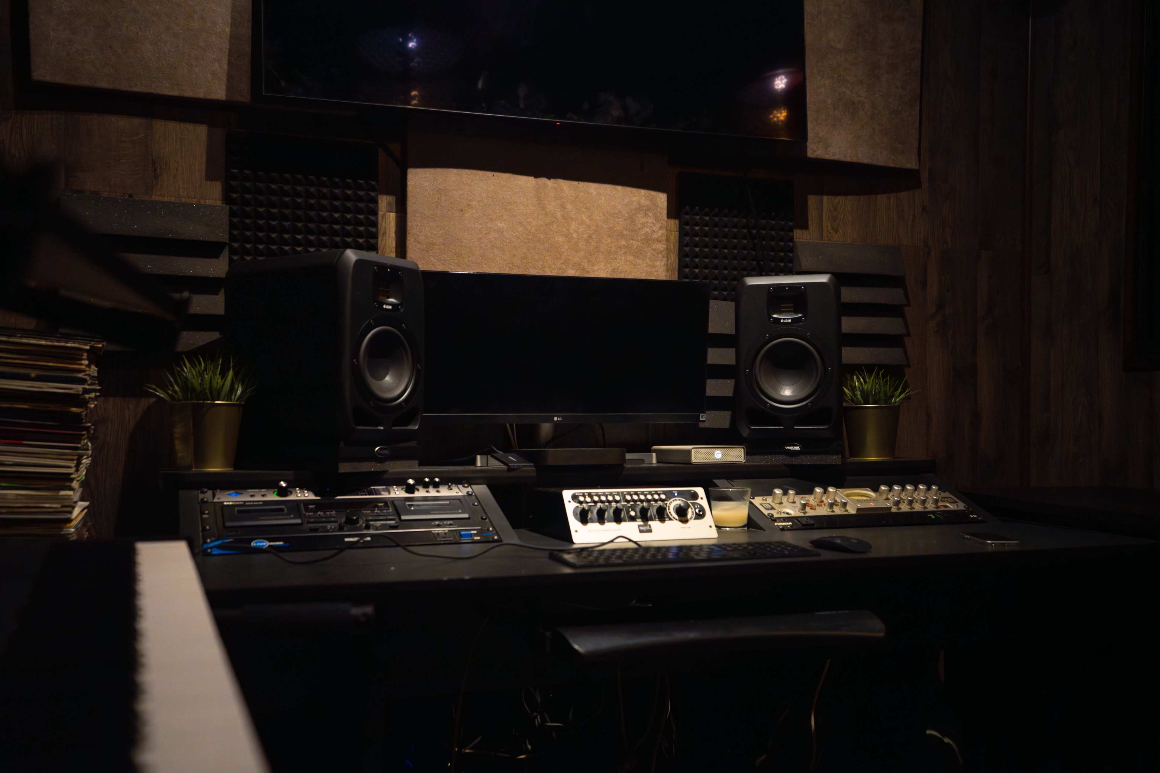 A music studio with an array of audio equipment, including speakers, a mixing console, and recording devices, arranged on a dark desk with a piano keyboard visible in the foreground.