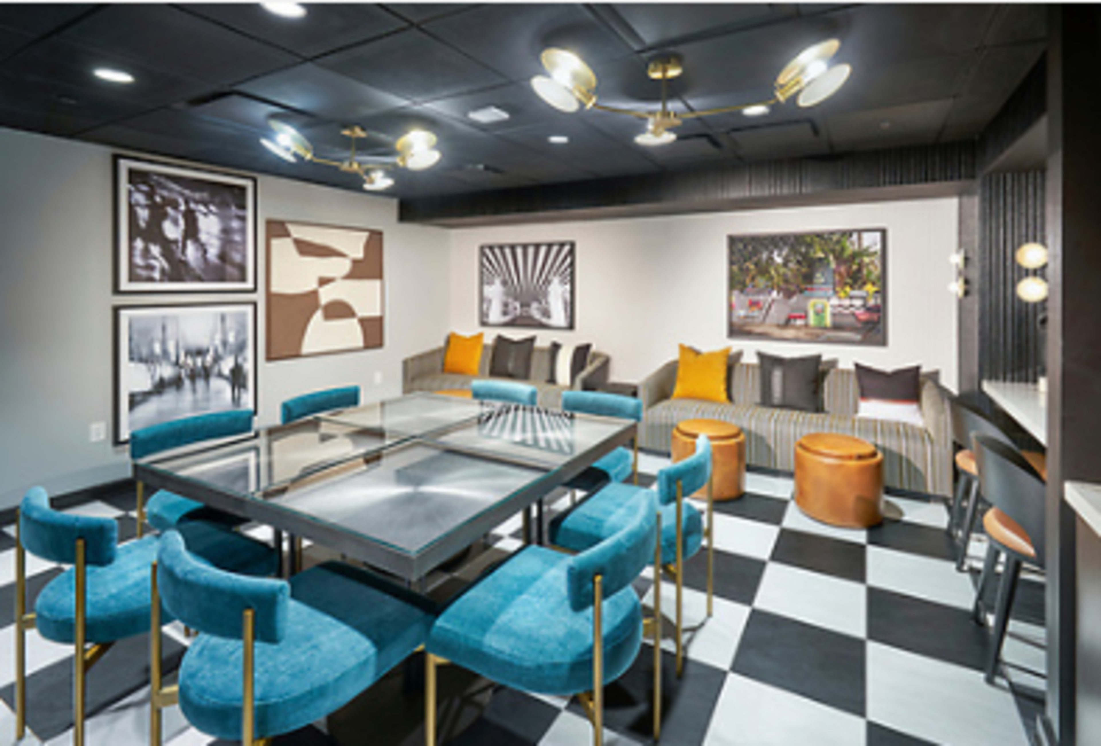 The image shows a modern lounge area with a glass table surrounded by blue chairs, artwork on the walls, and a mix of seating options, including a striped sofa and round stools.