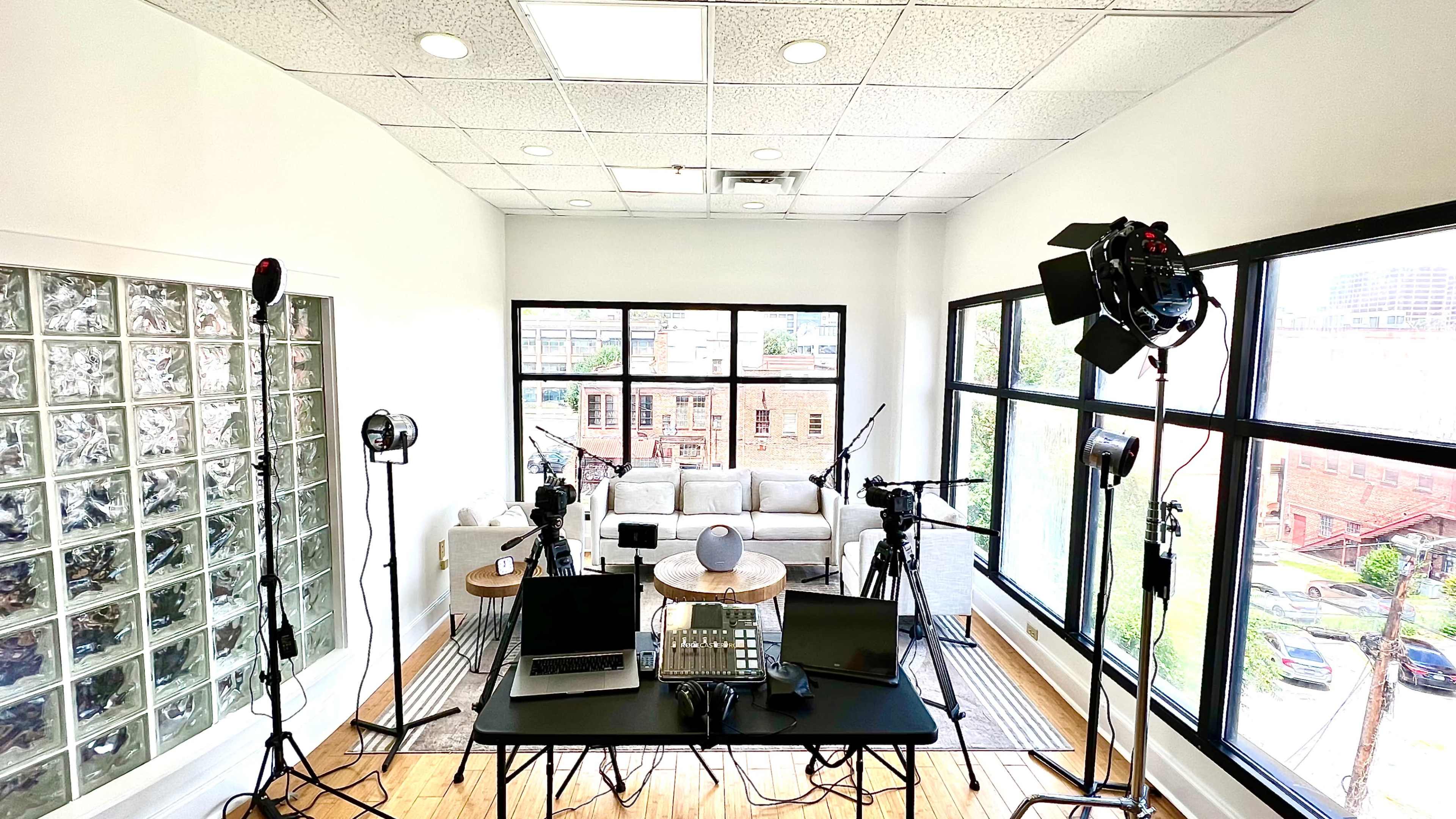 A modern studio setup with cameras, lights, and audio equipment arranged around a table in front of a large window with a view of the outside.