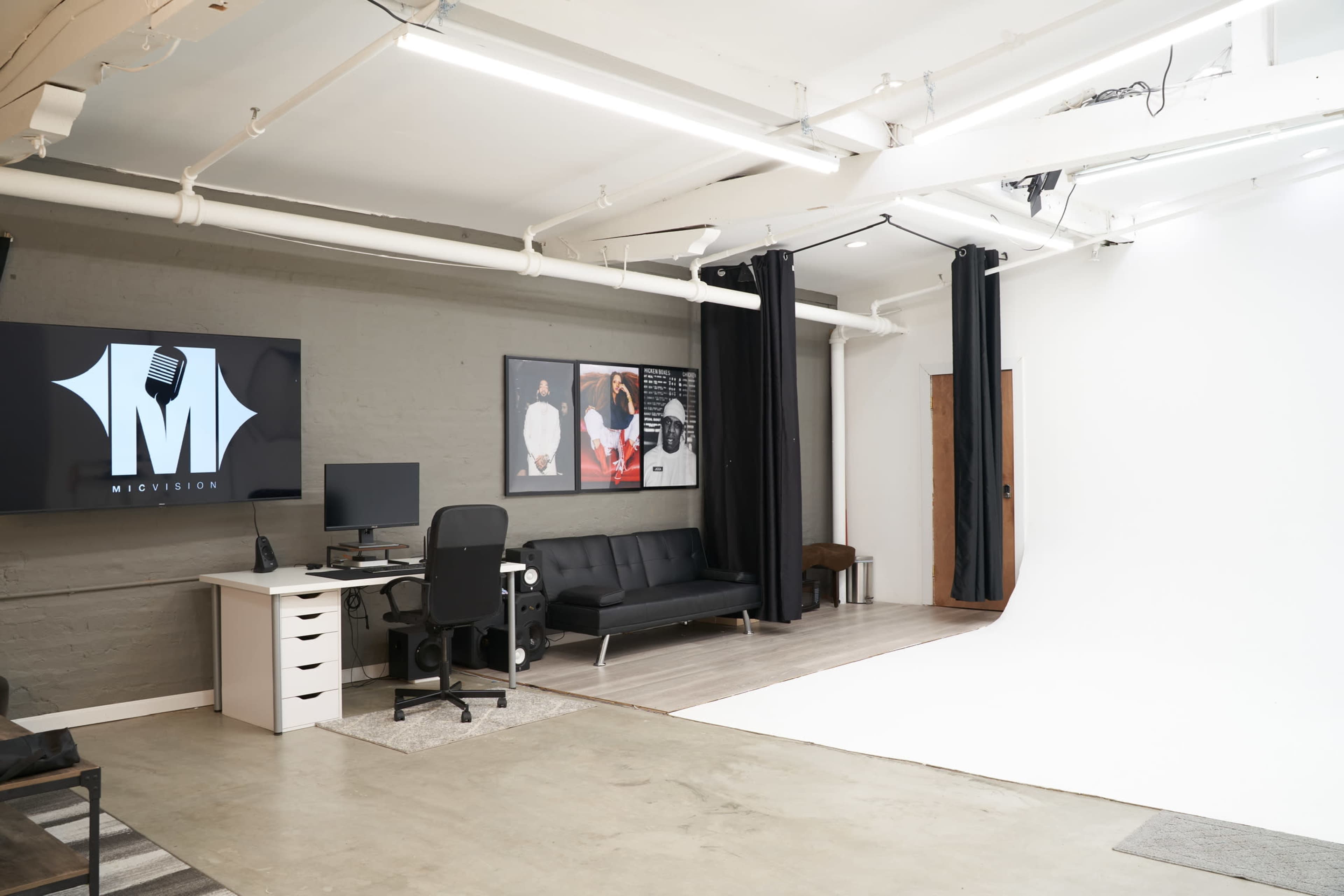 The image features a photography studio with a white backdrop, a desk with a computer, and a black couch, along with black curtains and framed photographs on the walls.