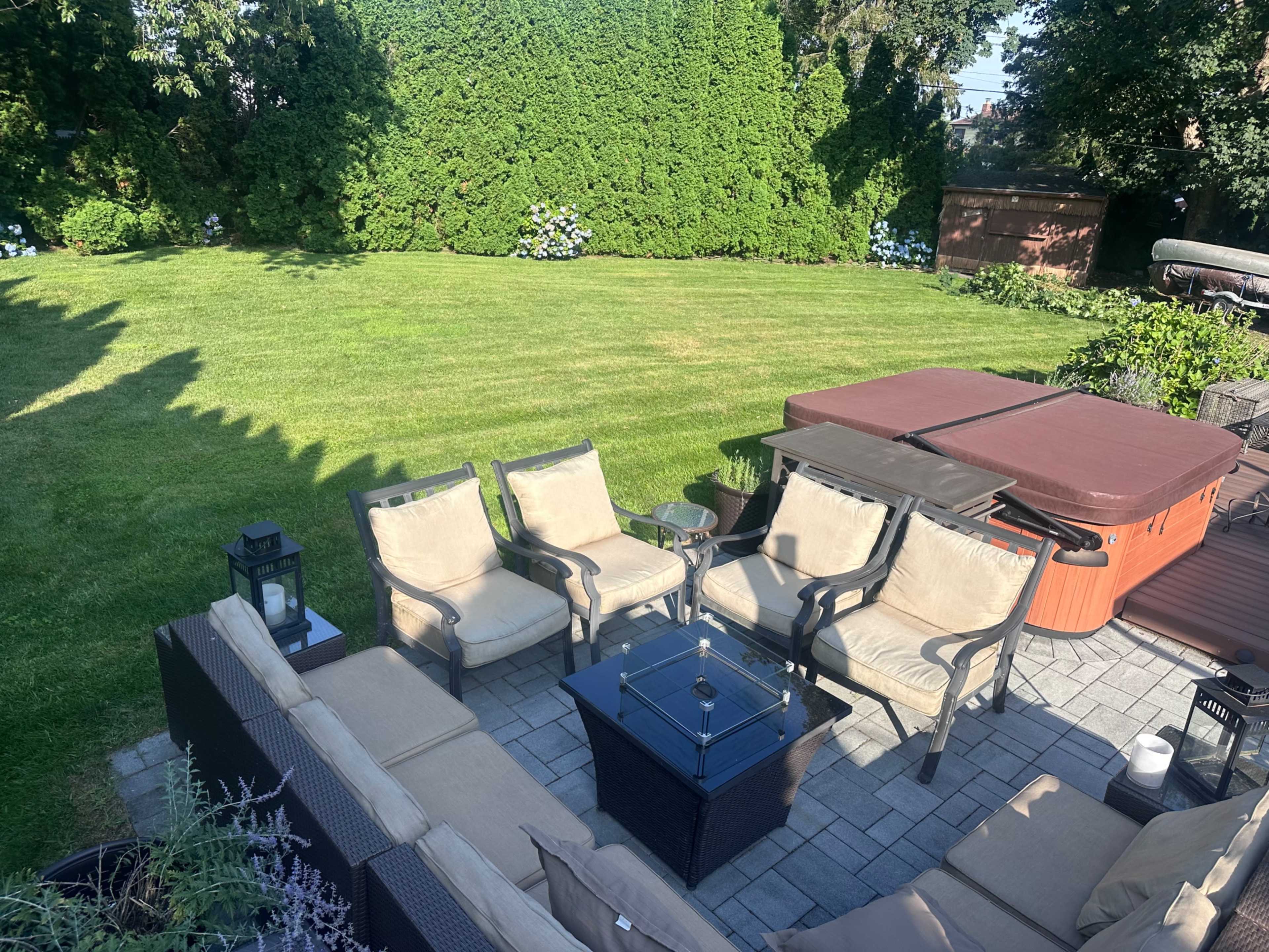 The image shows a landscaped backyard with a seating area, a table, and a hot tub surrounded by greenery.