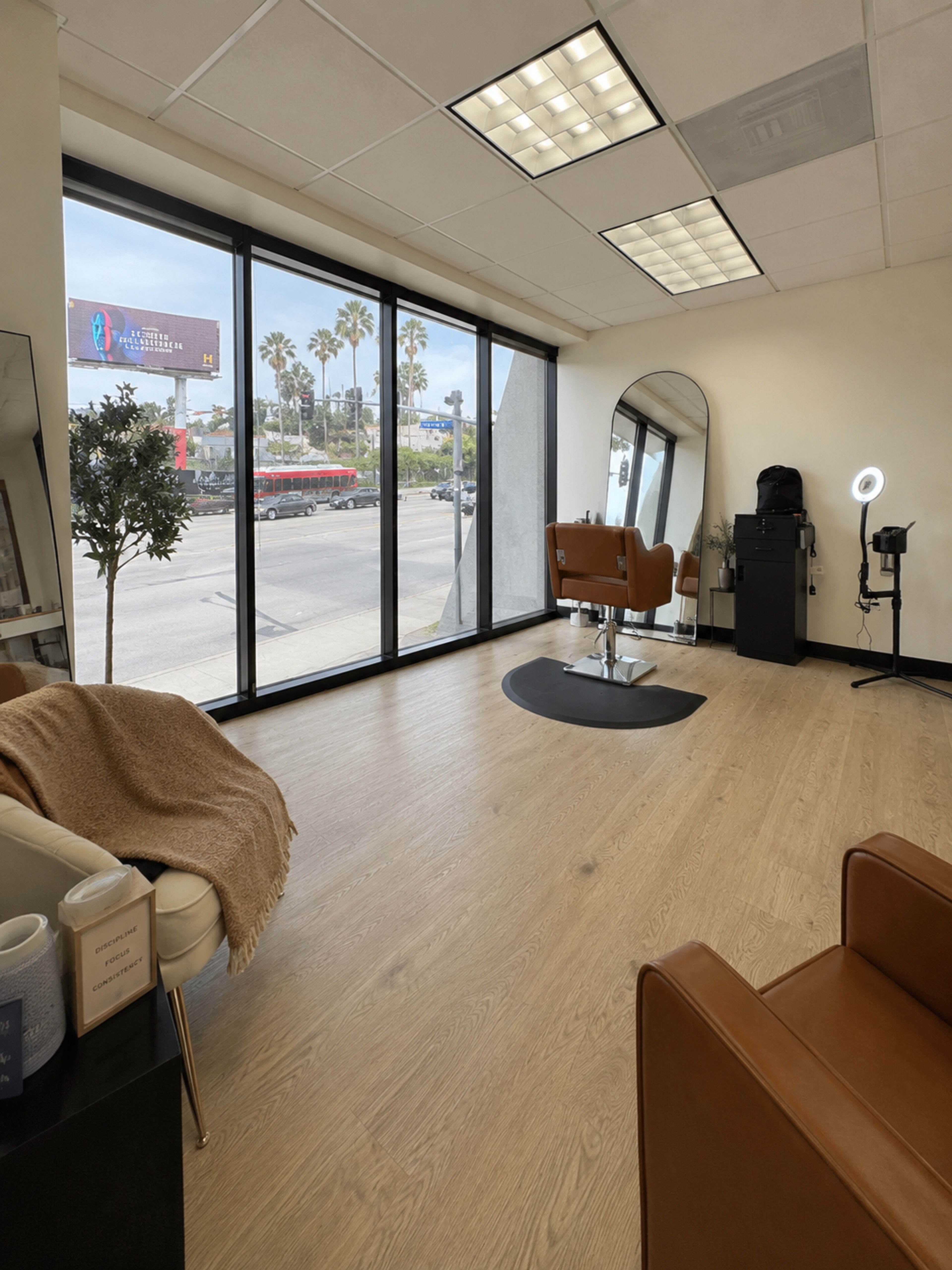 The image shows a spacious hair salon with large windows, a styling chair, a mirror, and minimal decor, overlooking a busy street.