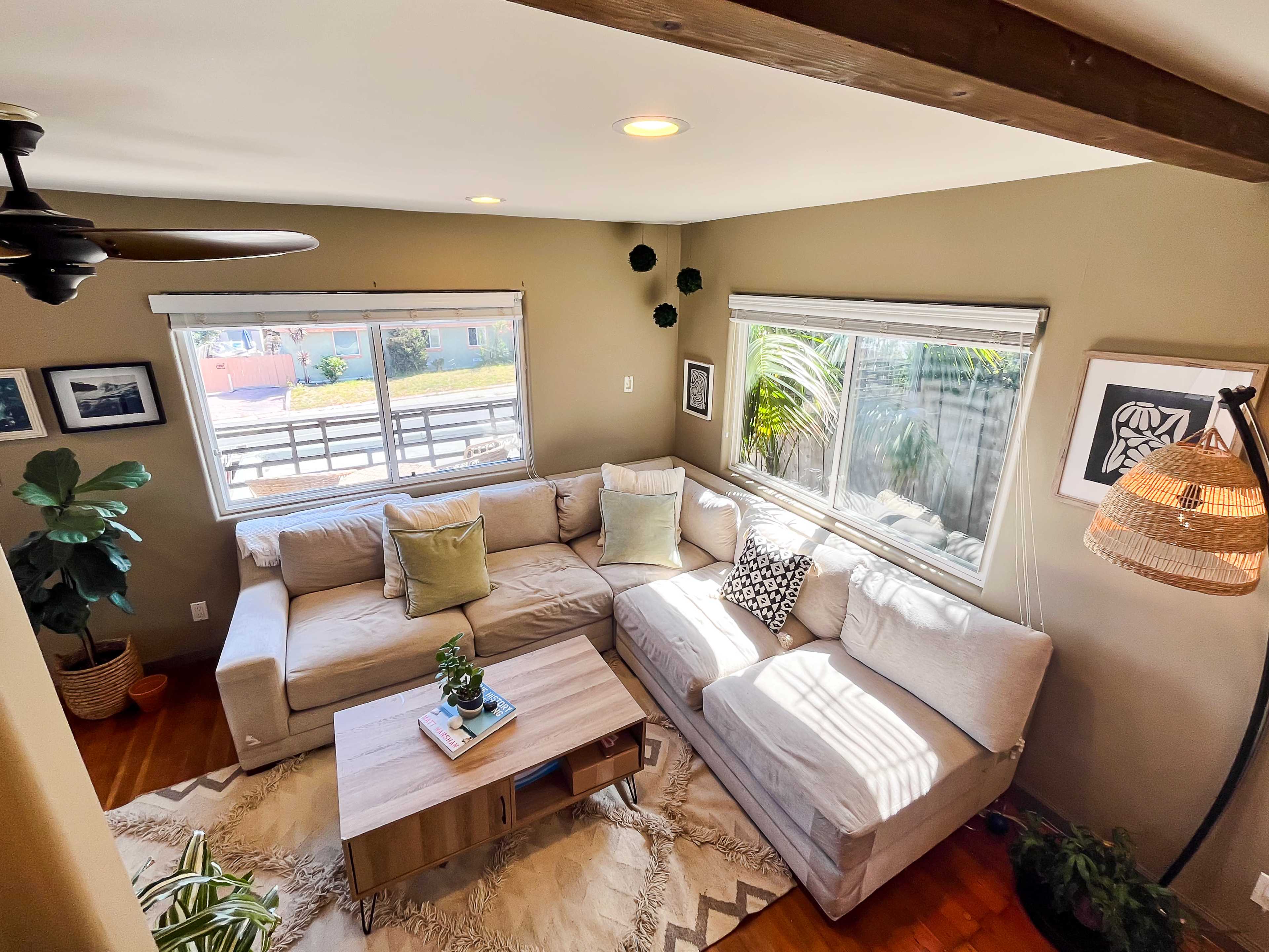 A well-lit living room features a sectional sofa, a wooden coffee table, and large windows with indoor plants.