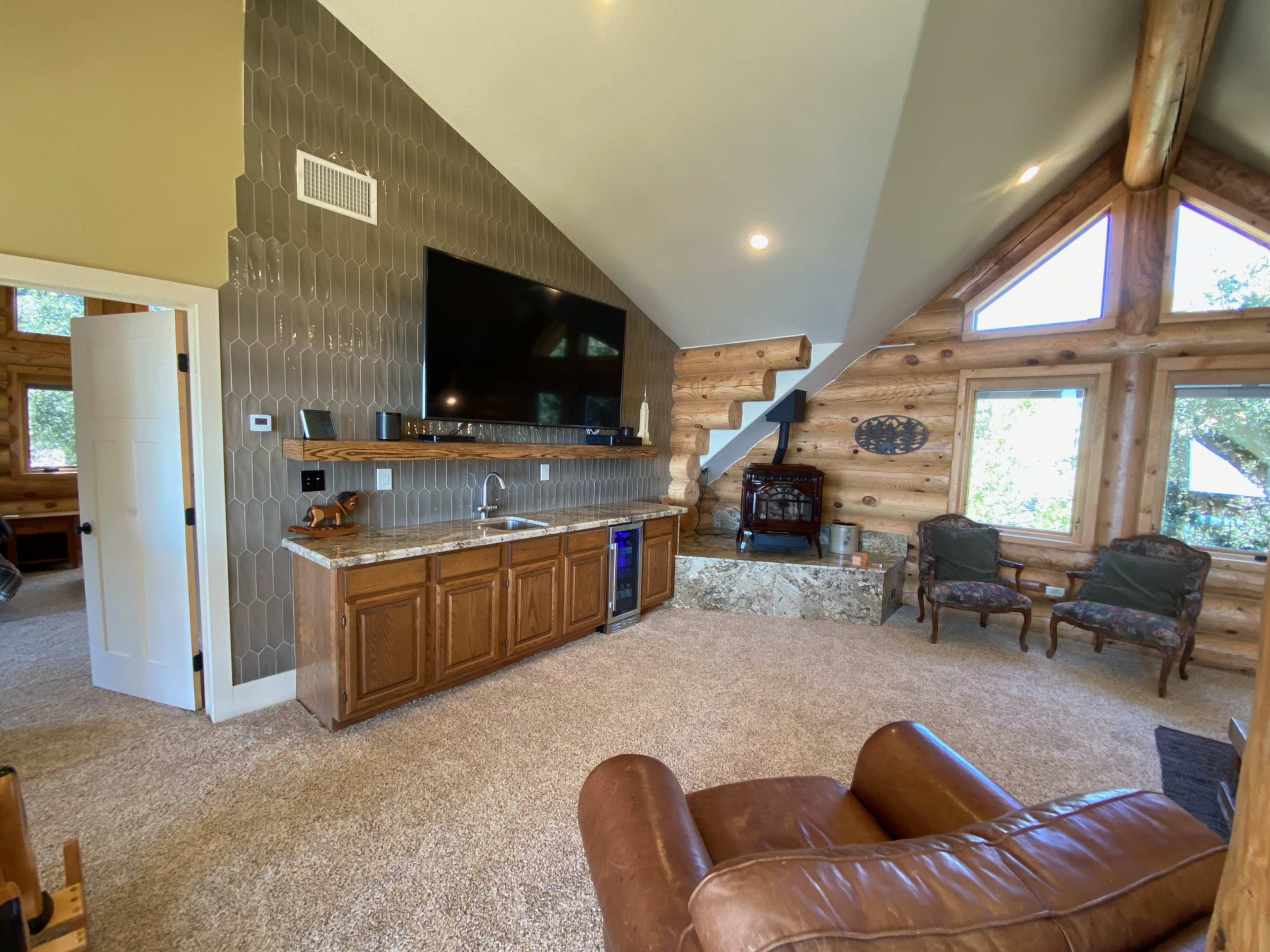 A cozy living area in a log cabin, featuring a stone fireplace, wooden shelves, and a large television mounted on the wall.