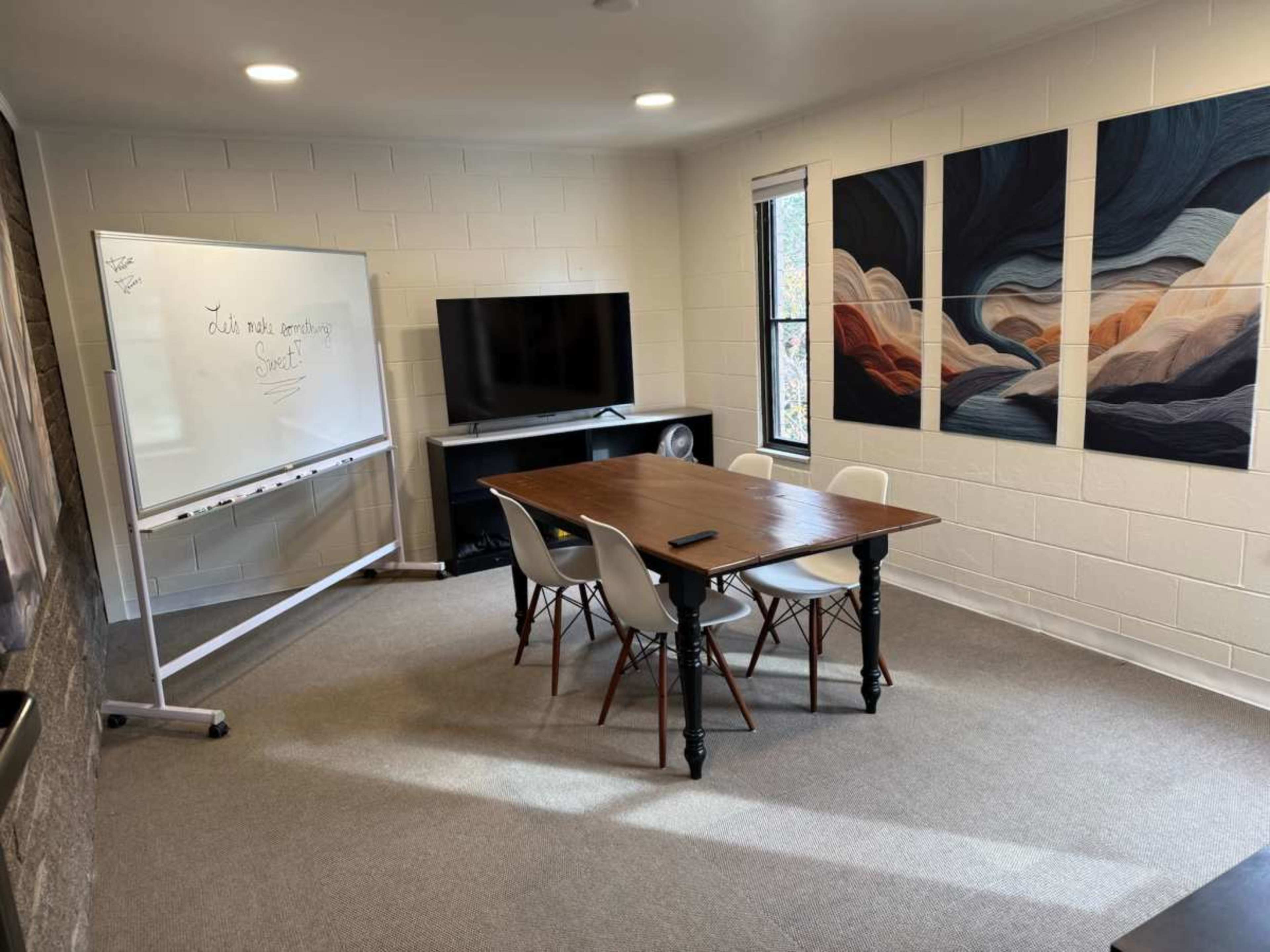The image shows a small meeting room featuring a wooden table with four chairs, a whiteboard, a television, and abstract art on the walls.