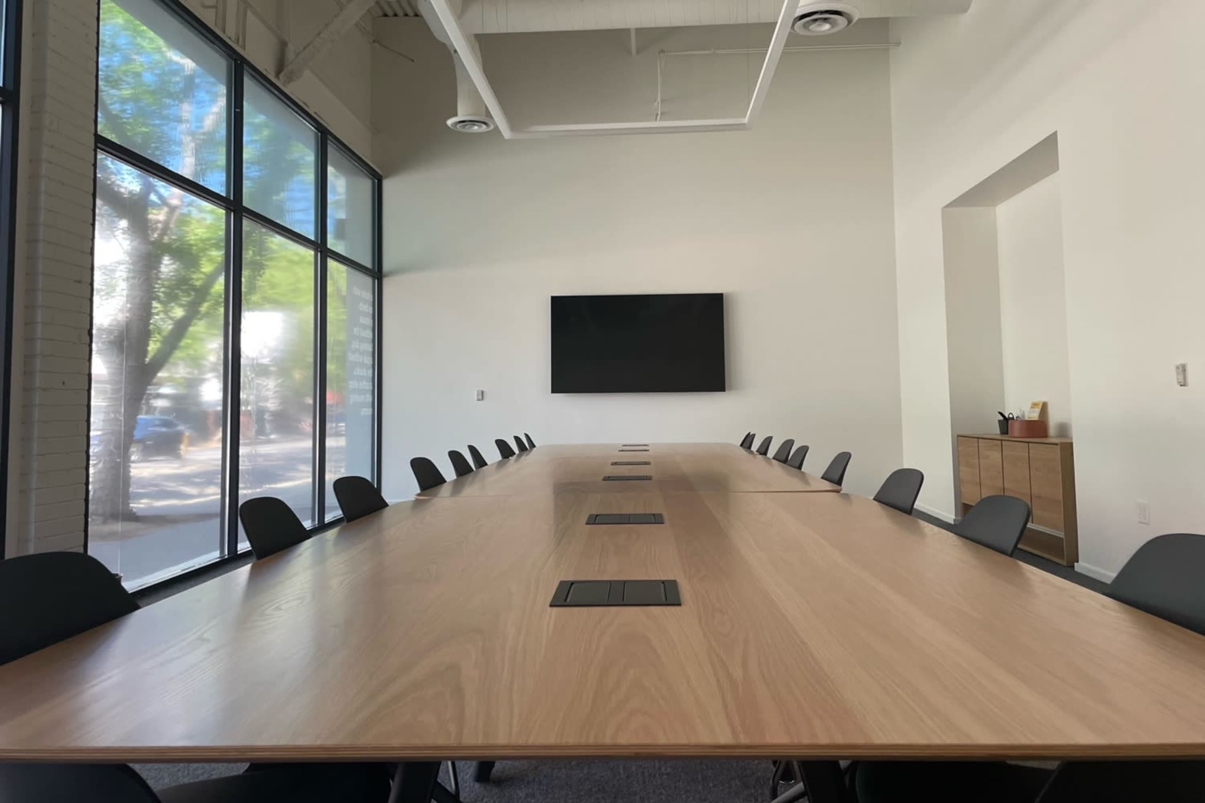 A large, modern conference room features a long wooden table with multiple seats and a wall-mounted screen at the front.