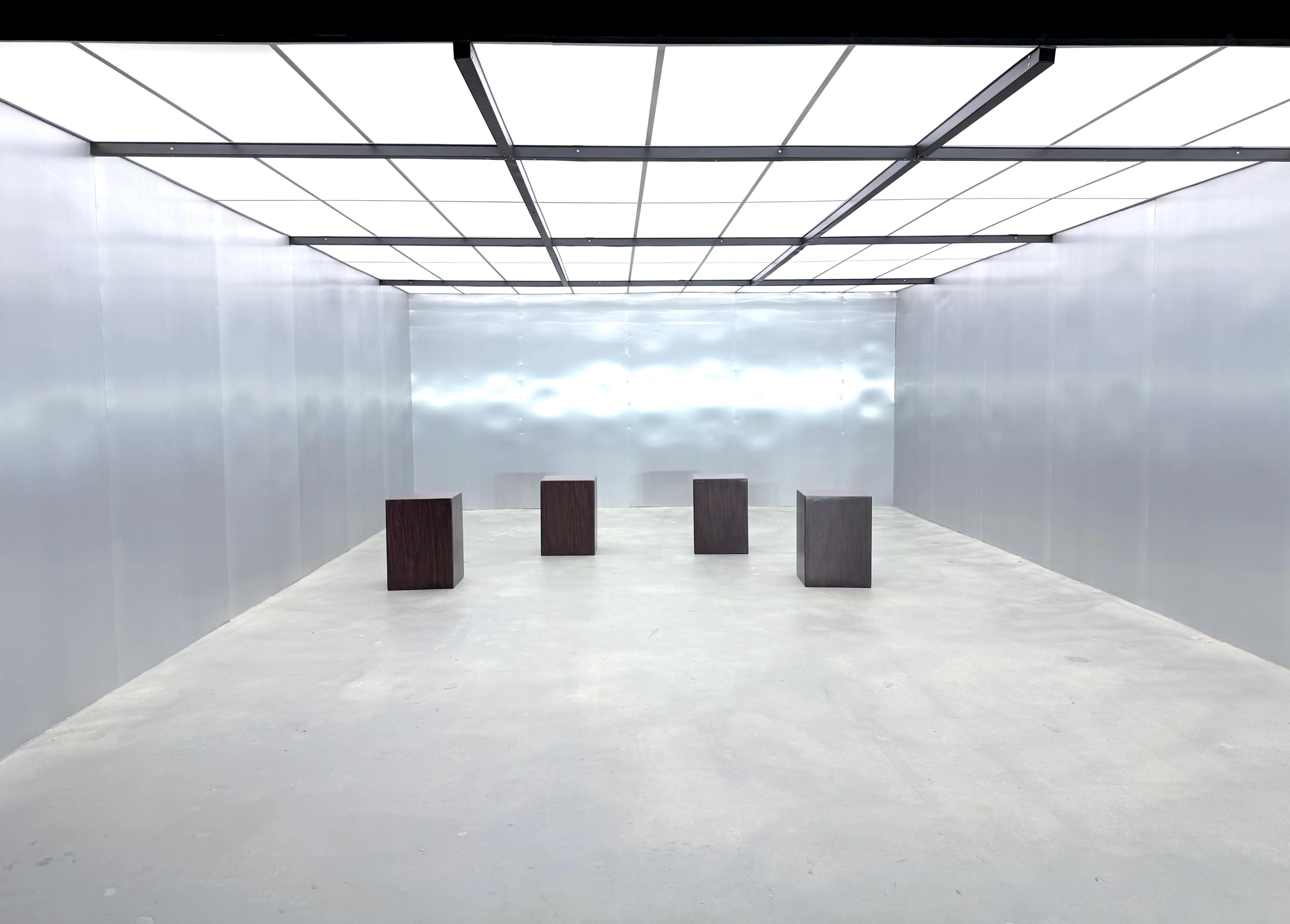 The image shows a minimalistic, empty room with four wooden blocks arranged in a line on a smooth concrete floor, surrounded by metallic walls and a bright, lit ceiling.