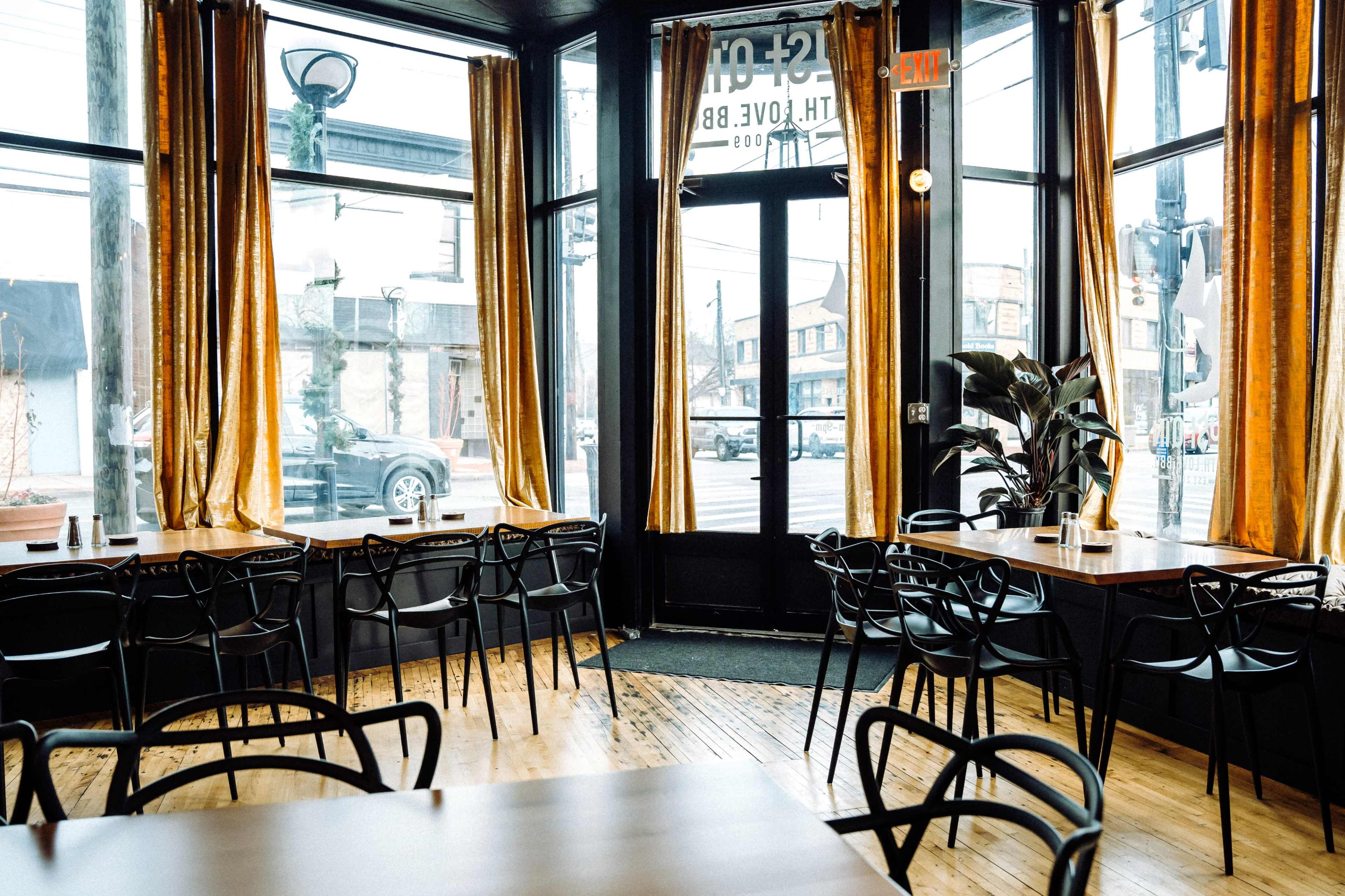 The interior of a cafe features wooden floors, tables set for dining, and large windows covered with golden curtains, allowing natural light to fill the space.