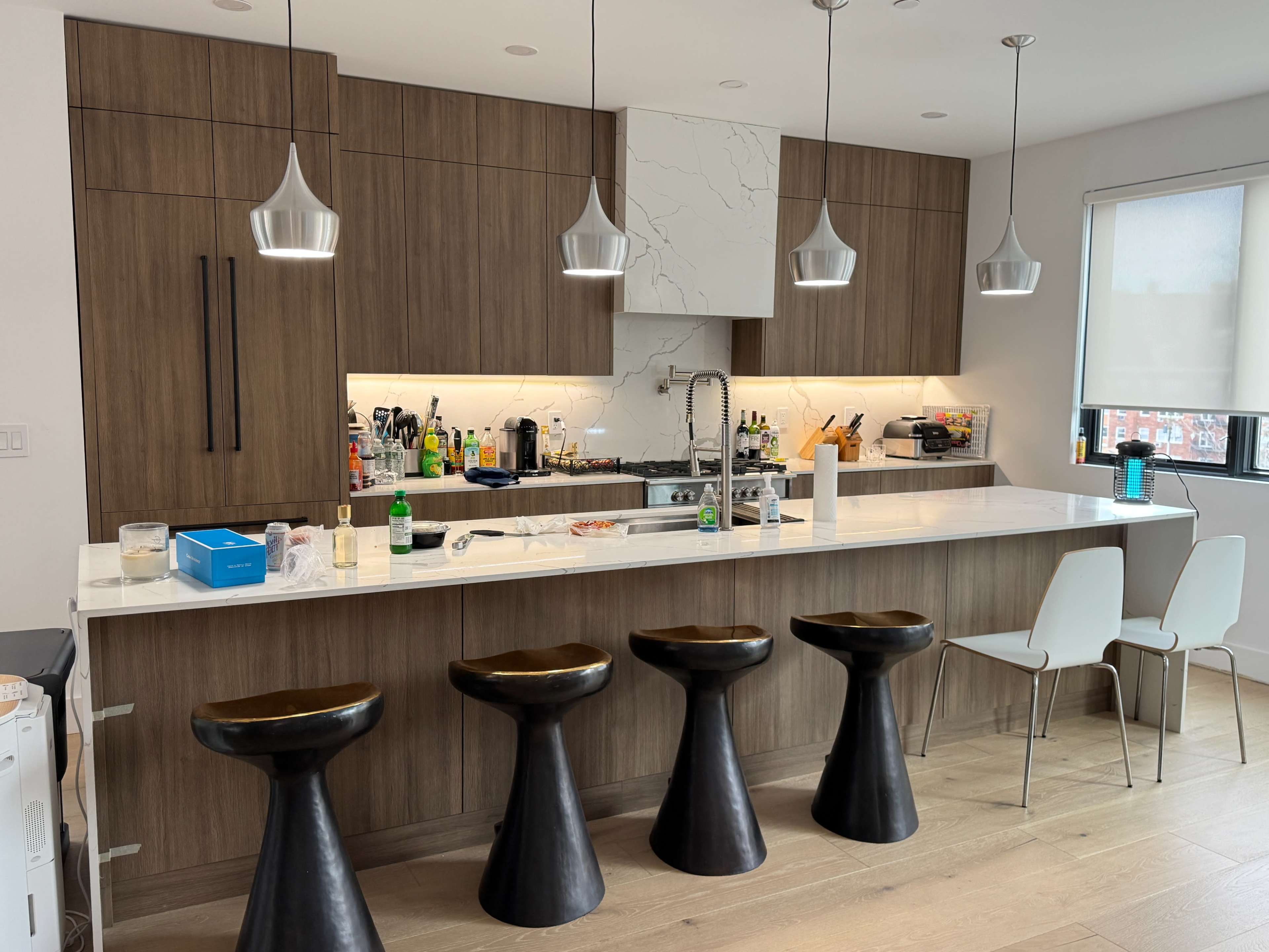 The image shows a modern kitchen with wooden cabinets, a marble countertop, and four black stools lined up at a bar.