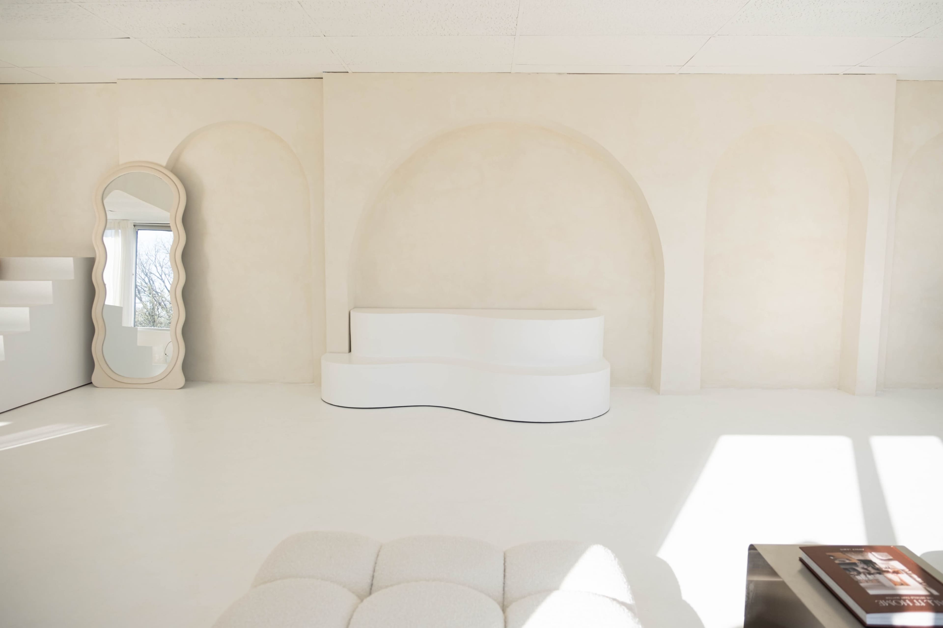 The image shows a minimalist interior with curved architectural features, a white sculptural bench, and a large ornate mirror against a light-colored wall.