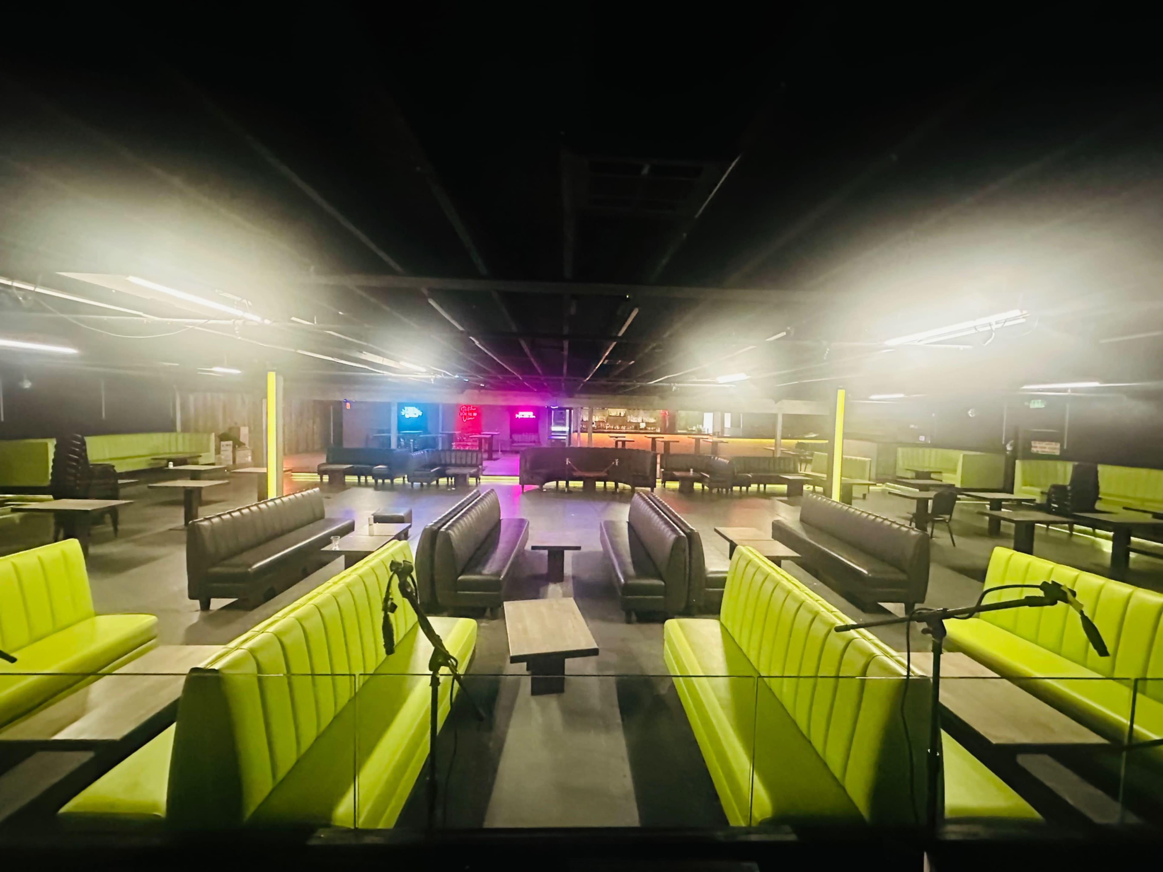 The image shows an empty indoor venue with green upholstered seating, dim lighting, and a bar area in the background.