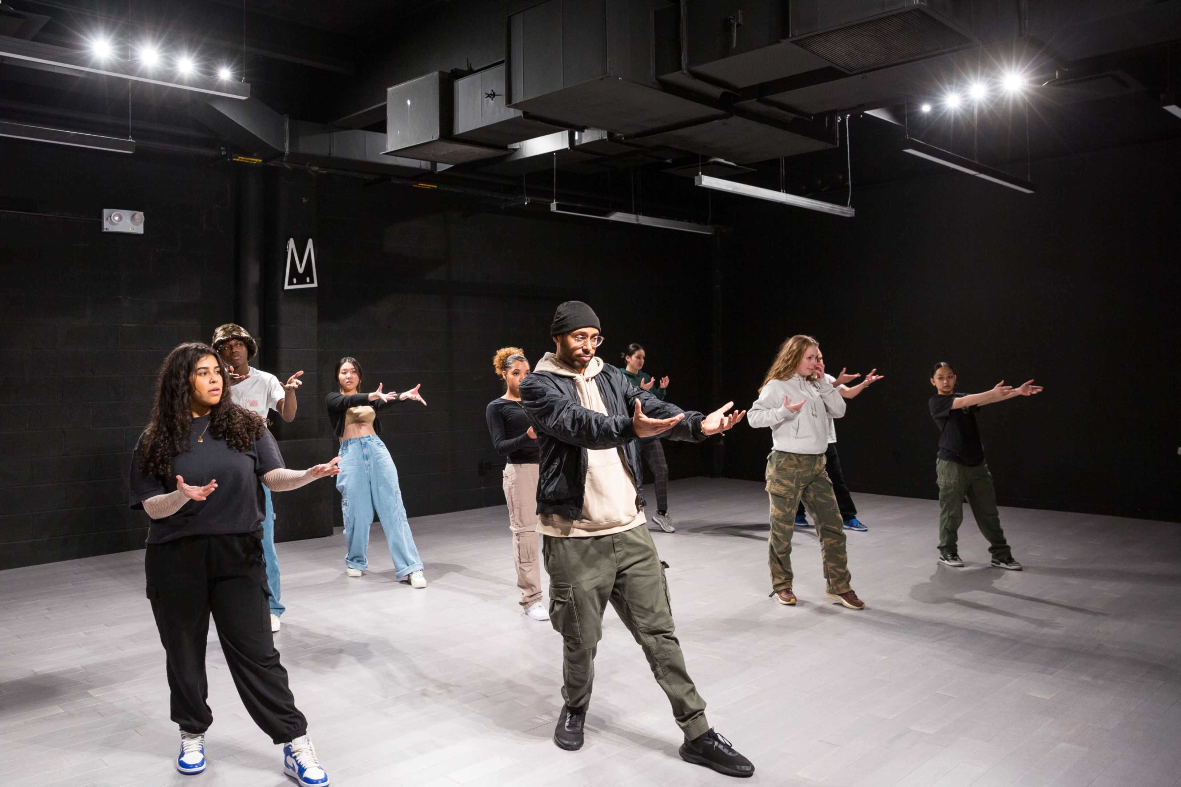 A group of dancers is practicing choreography in a studio with a dark background and bright overhead lights.