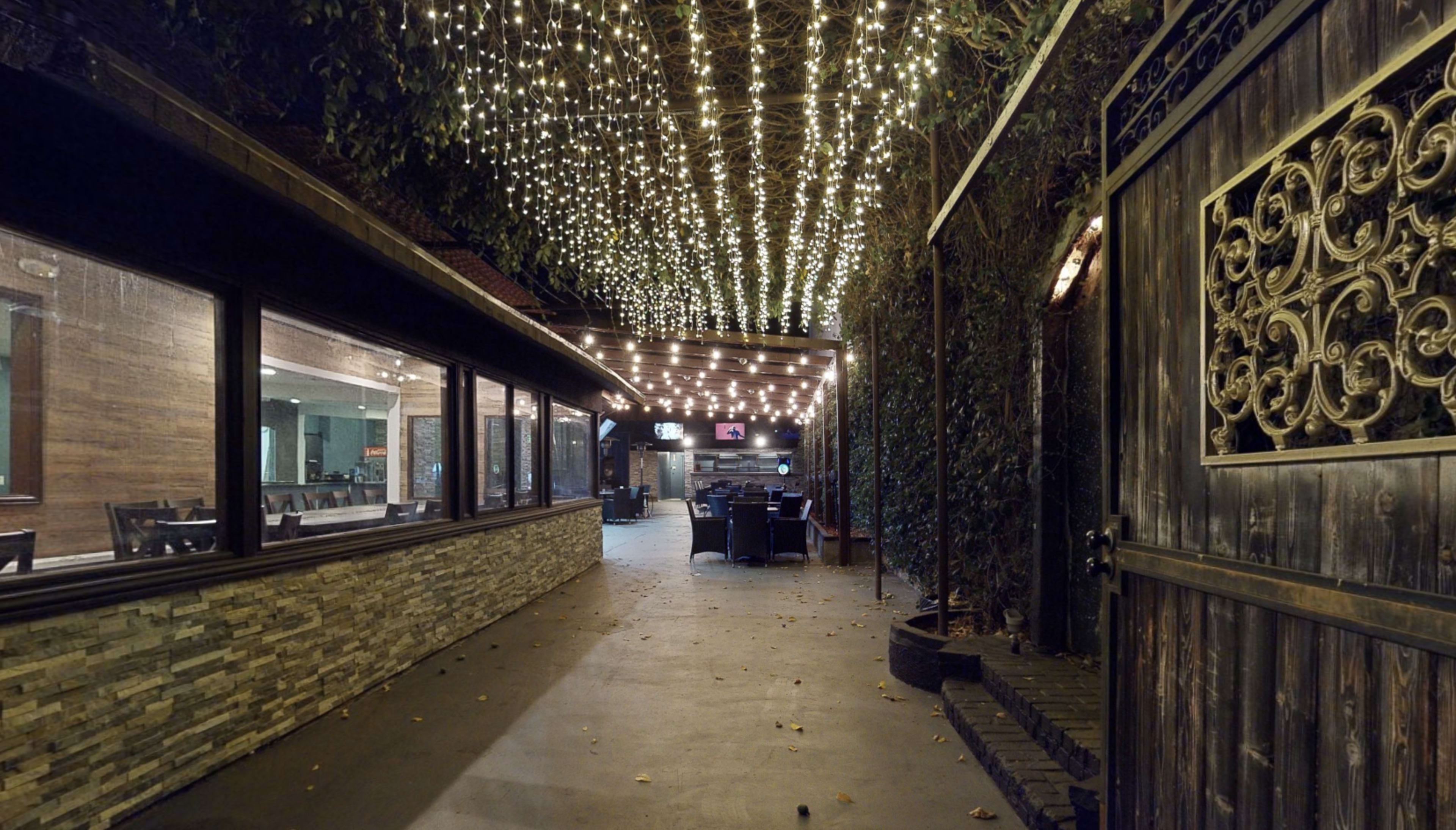 A pathway lined with greenery leads to an outdoor dining area illuminated by string lights overhead.