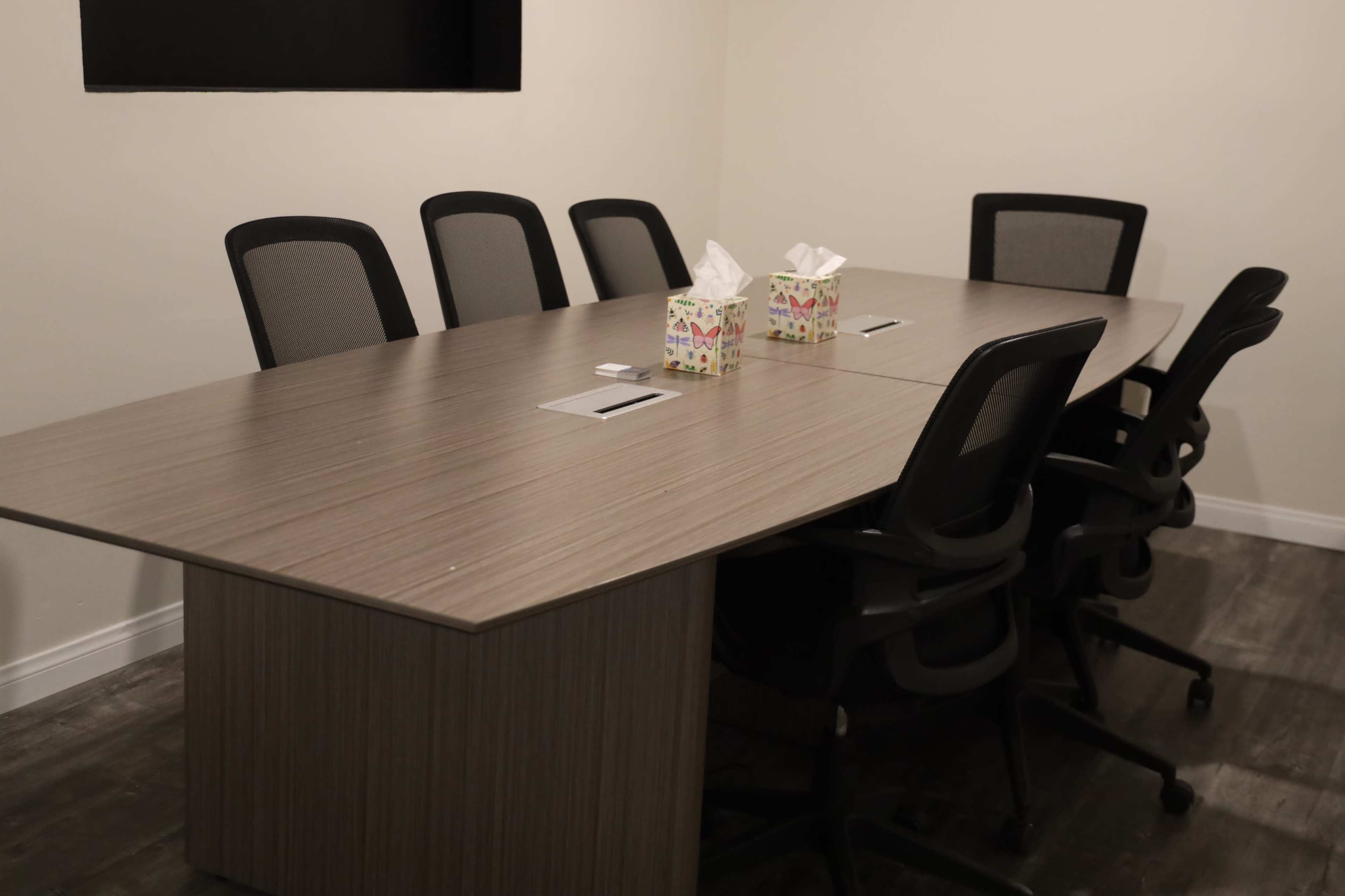 The image shows a conference room with a long wooden table and several black ergonomic chairs arranged around it, featuring two boxes of tissues on the table.