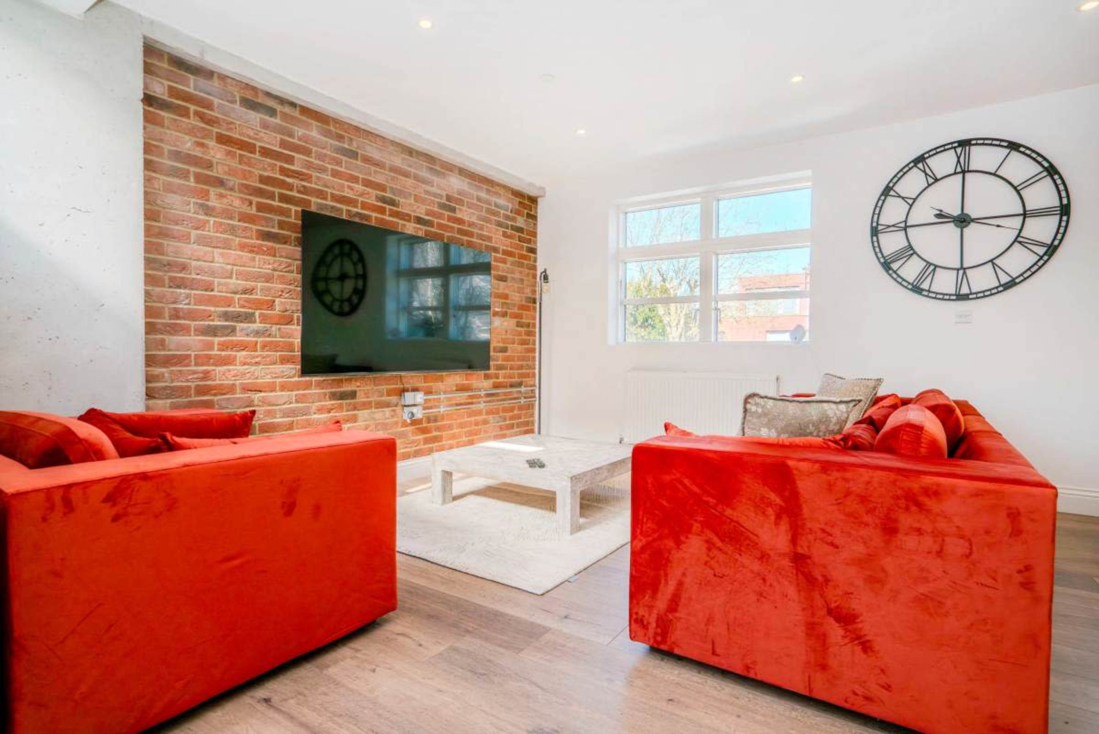 The image depicts a modern living room featuring a brick wall, a large wall clock, a flat-screen TV, and two red sofas facing a coffee table.