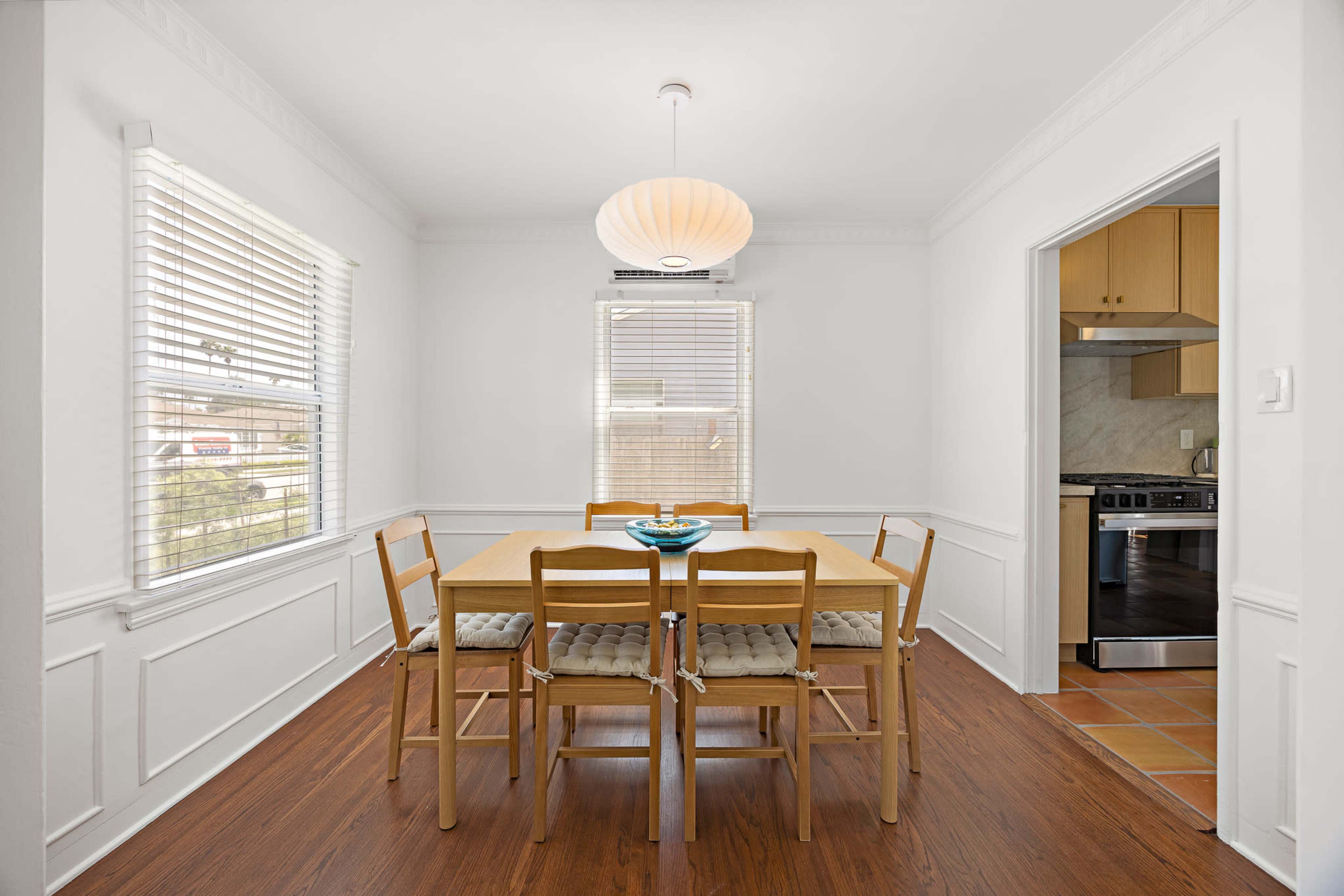 A simple dining area features a square wooden table surrounded by six chairs, with a pendant light above and a doorway leading to the kitchen.