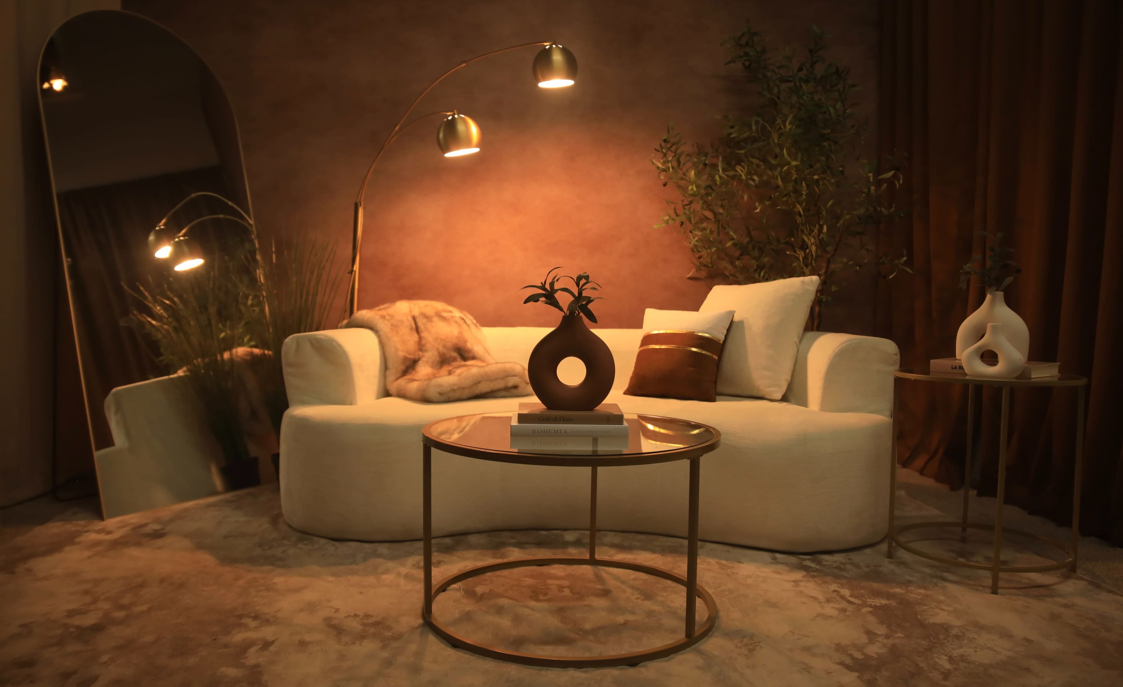 The image shows a cozy living room setting featuring a white curved sofa, a round glass coffee table, decorative vases, and warm lighting.