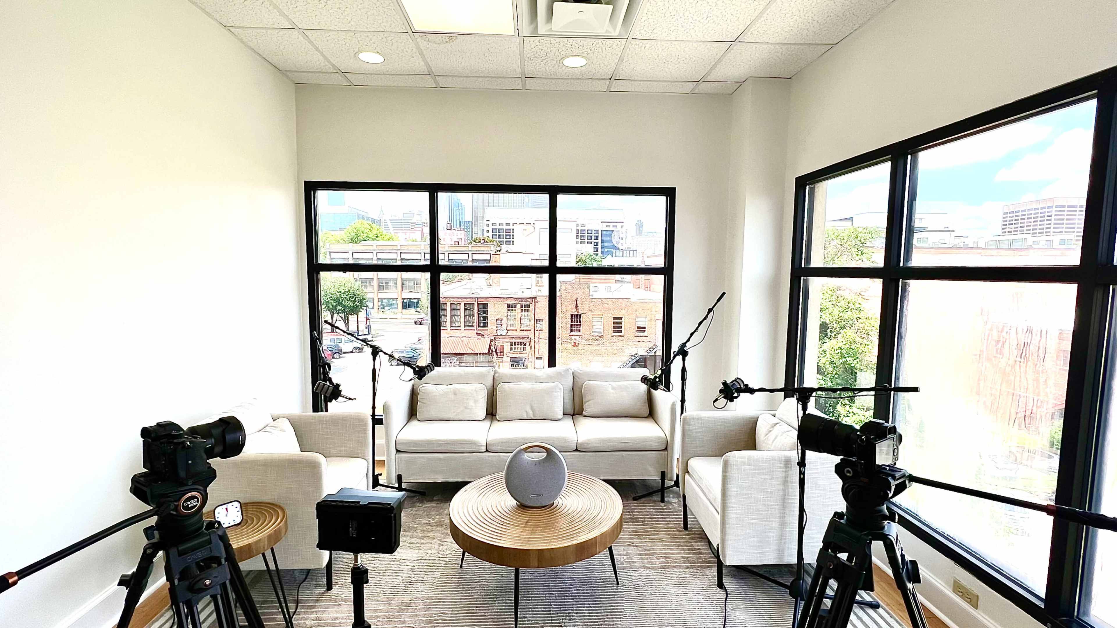 A modern, well-lit room with a set of recording cameras and microphones facing a circular table surrounded by white couches, overlooking a cityscape through large windows.