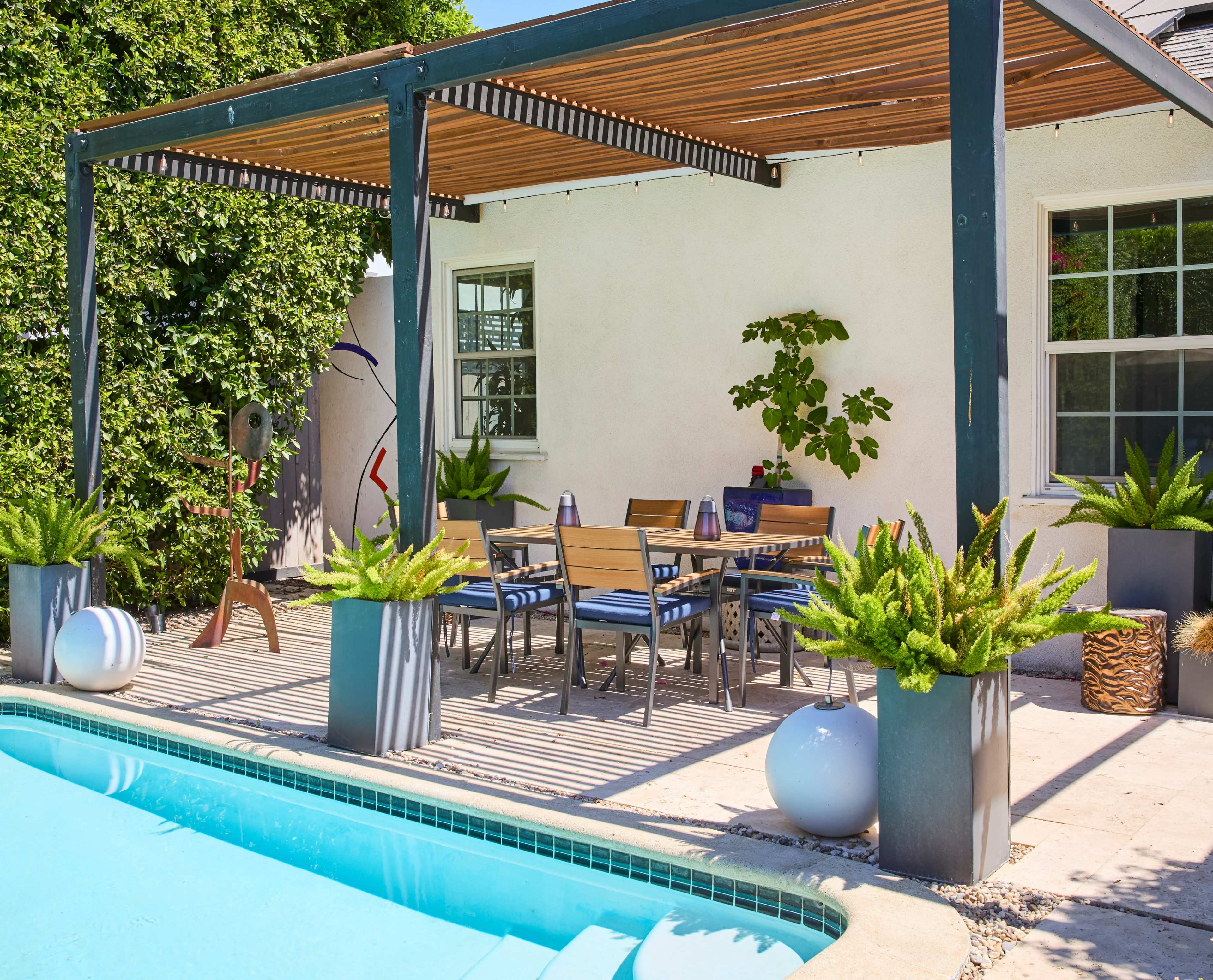A patio area with a dining table and chairs under a wooden pergola, alongside a swimming pool and various potted plants.