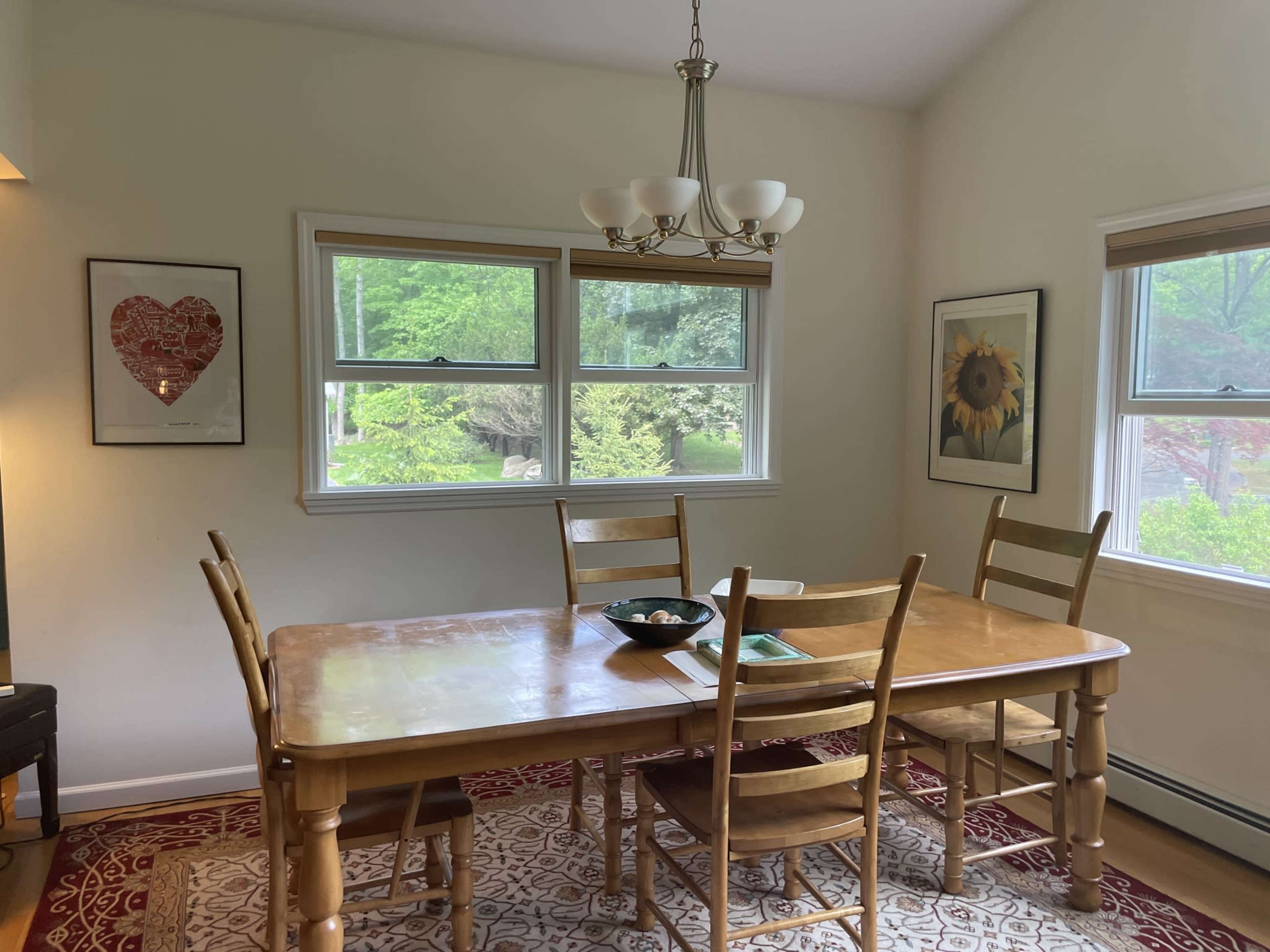 A wooden dining table with six chairs is set in a well-lit room featuring wall art and windows that overlook a garden.