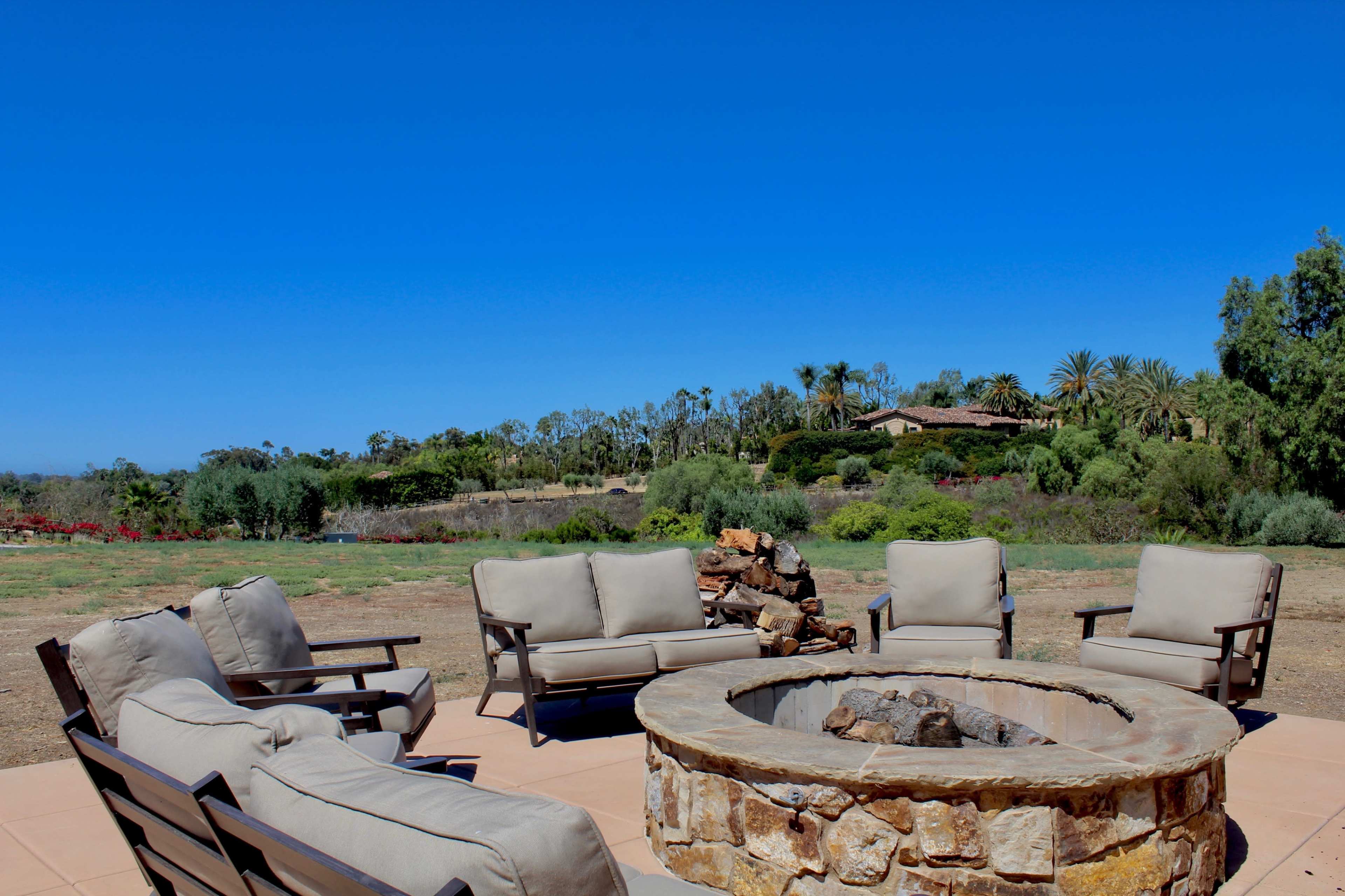 A stone fire pit surrounded by several cushioned chairs overlooks a spacious outdoor landscape with trees and distant buildings under a clear blue sky.