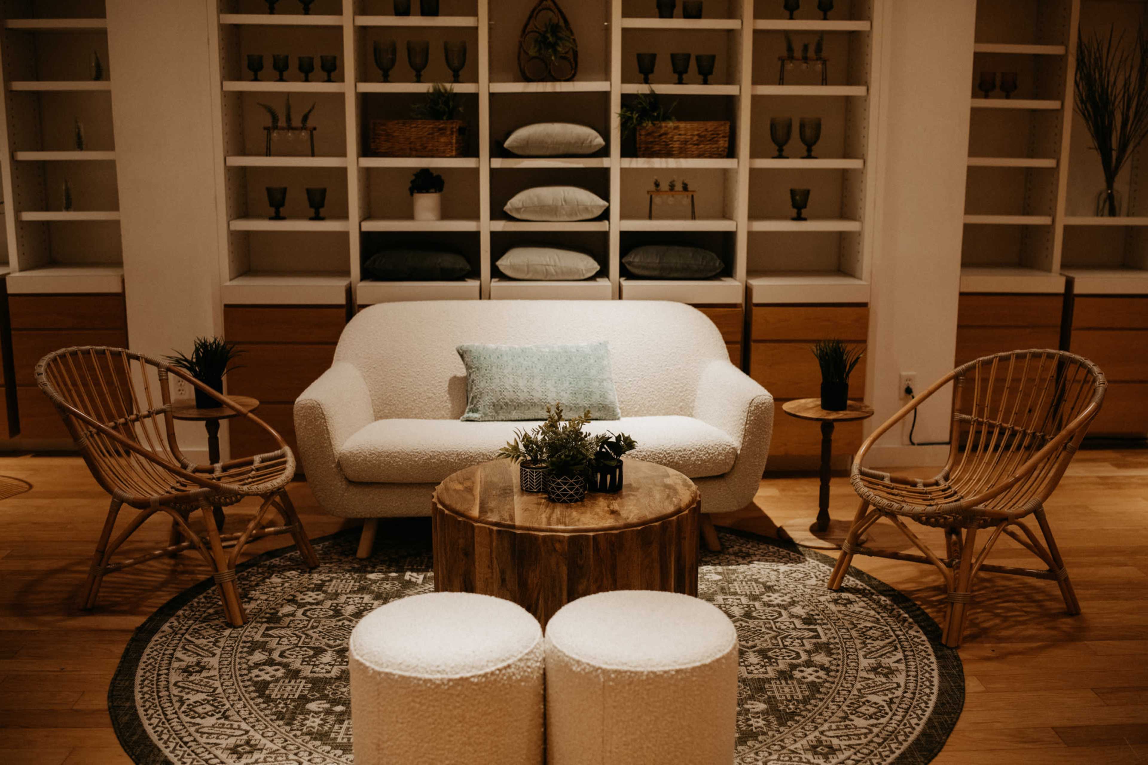A modern living room features a white sofa, decorative plants, a wooden coffee table, and woven chairs arranged around a round rug.