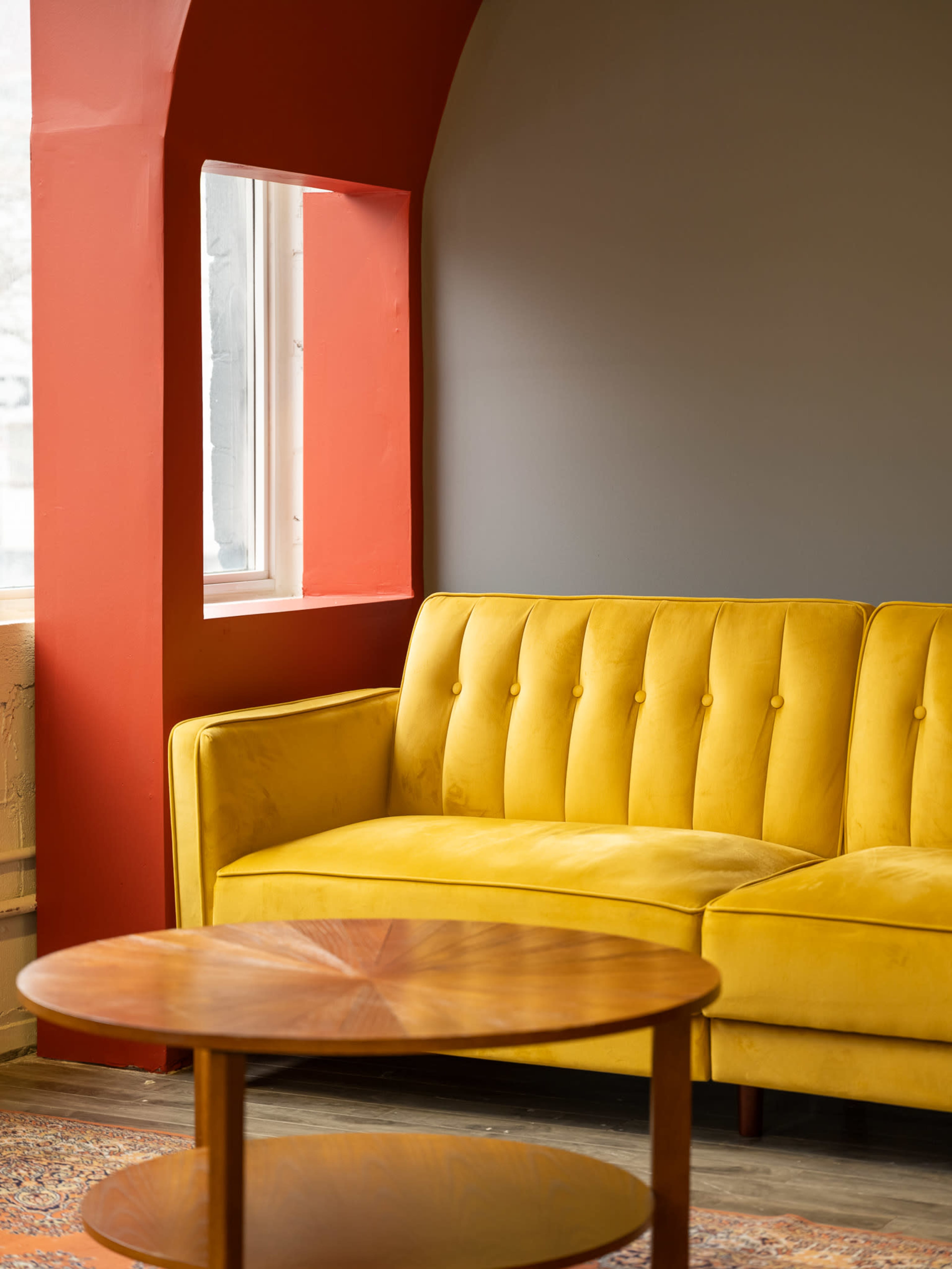 A yellow tufted couch is positioned next to a window, with a round wooden coffee table in front of it, against a gray wall accented by a red arch.