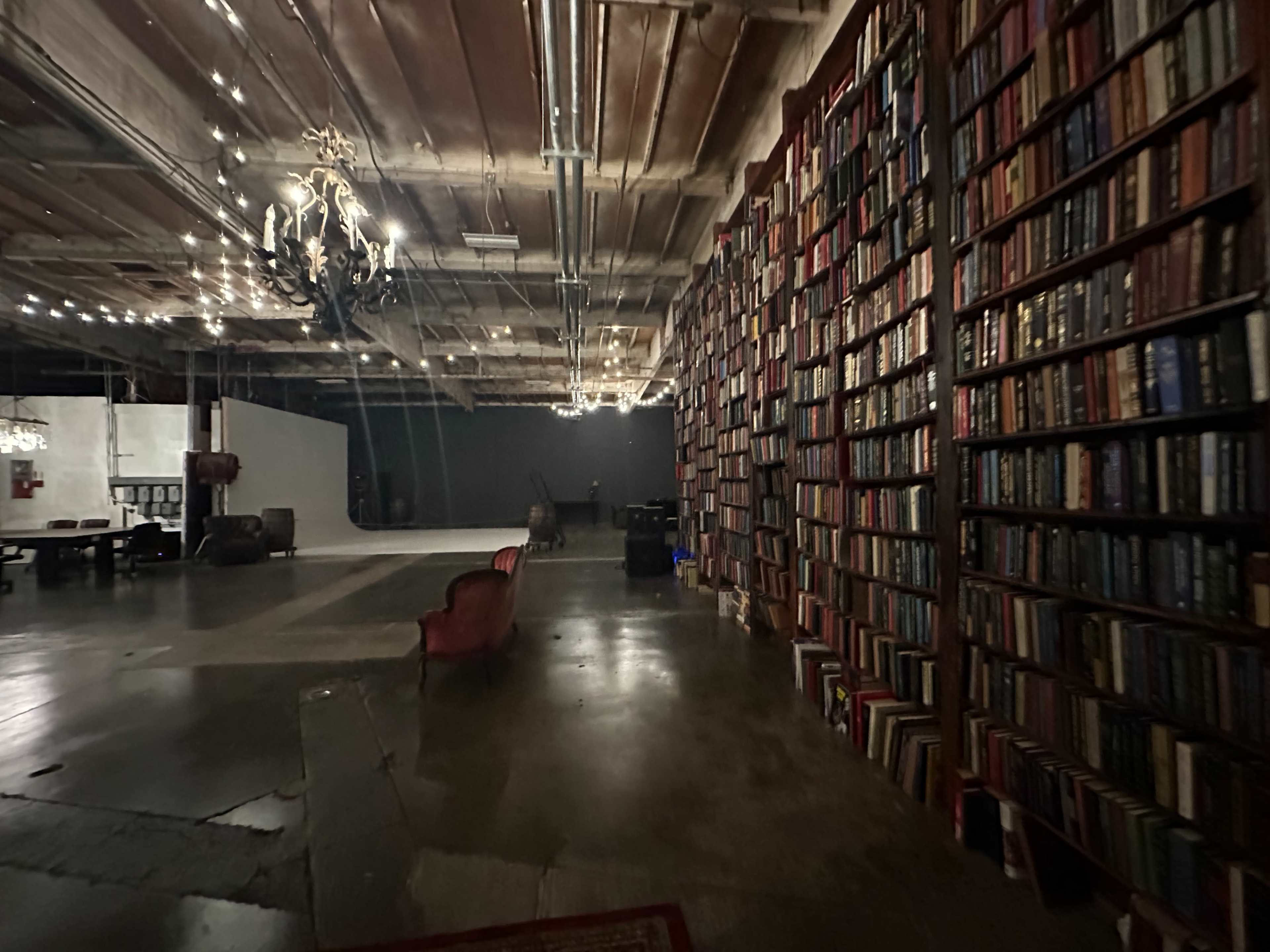 A dimly lit room with a tall wall of books and a red couch in the foreground, illuminated by overhead lights and a chandelier.