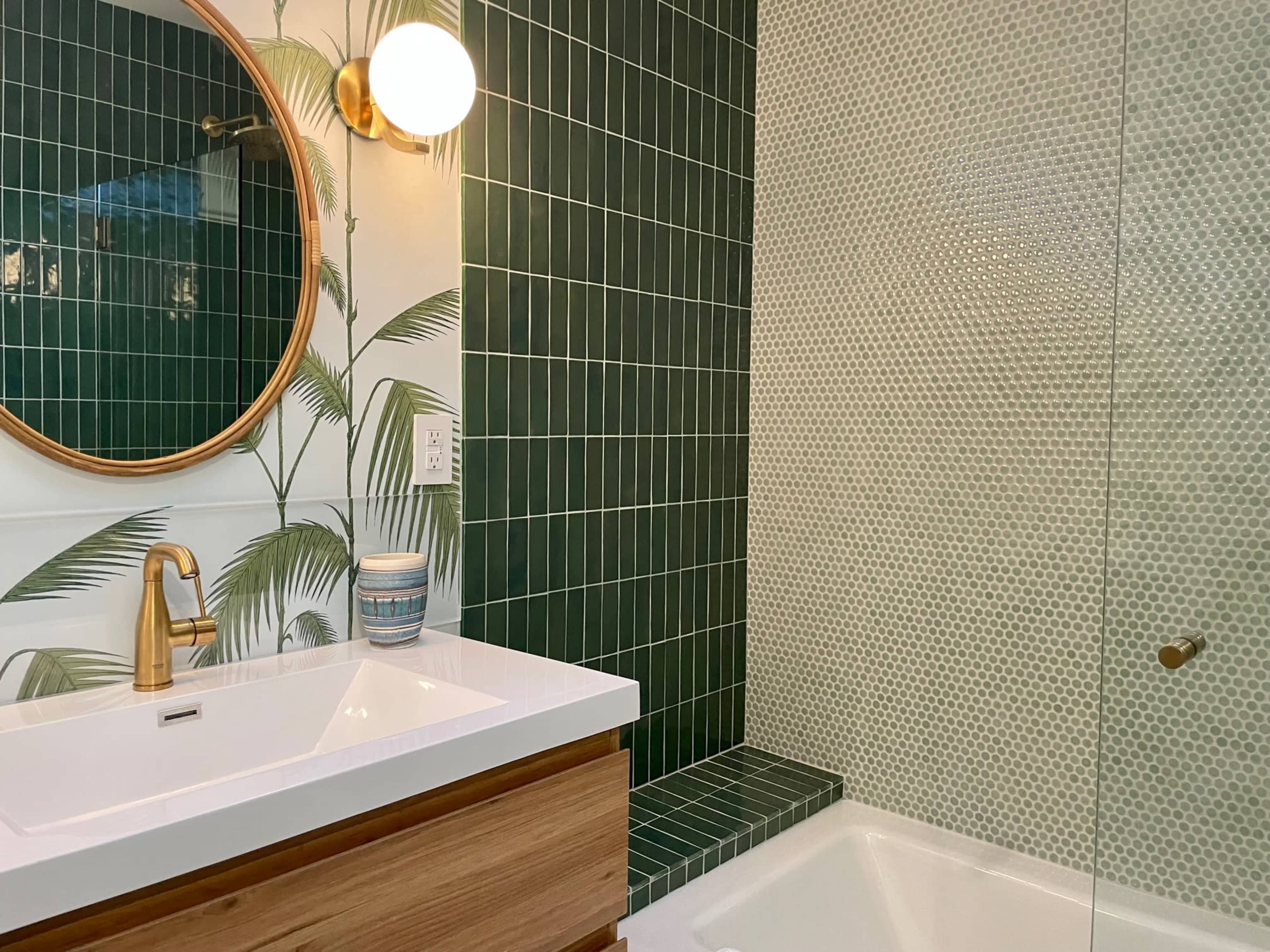The image shows a modern bathroom featuring a round mirror, a white sink with a wooden cabinet, and green tiled walls, with one section displaying hexagonal tiles and a glass shower enclosure.