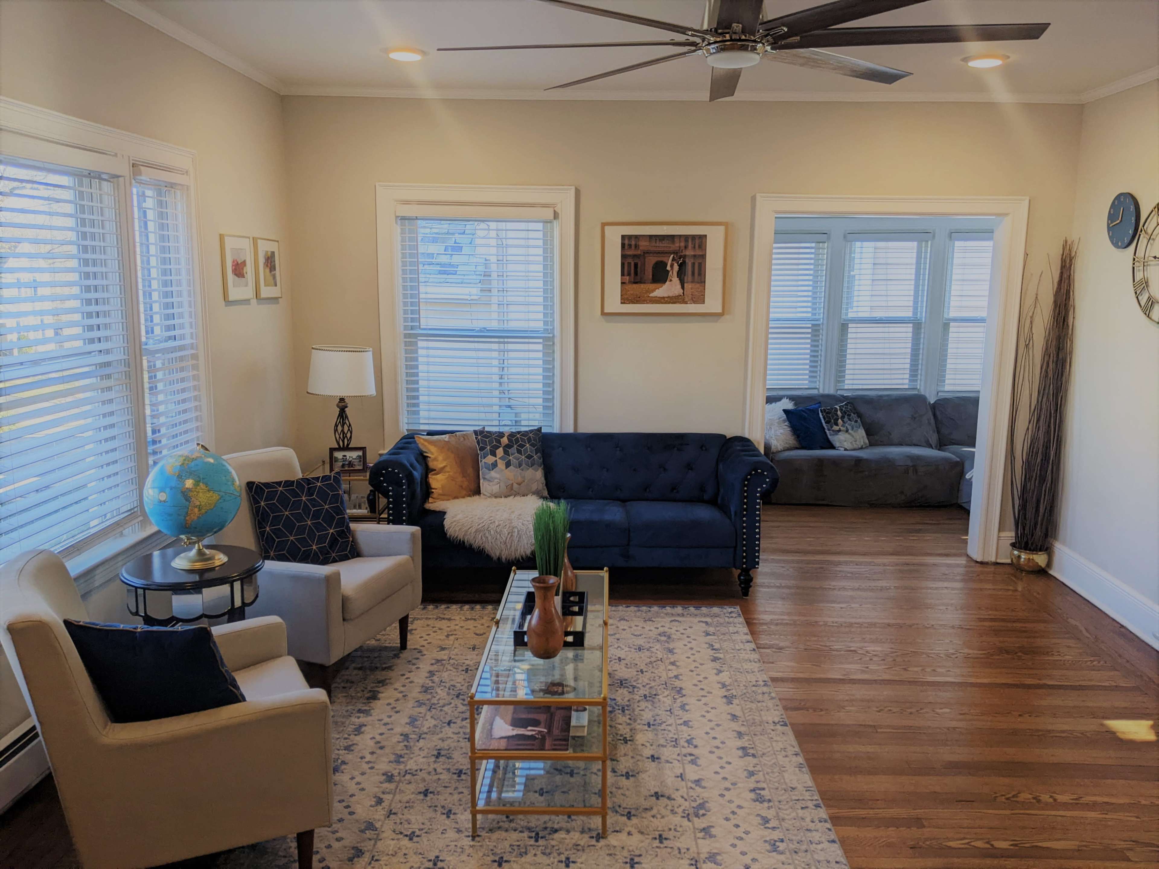 The image shows a bright living room featuring a blue couch, beige armchairs, a coffee table, and large windows allowing natural light to flow into the space.