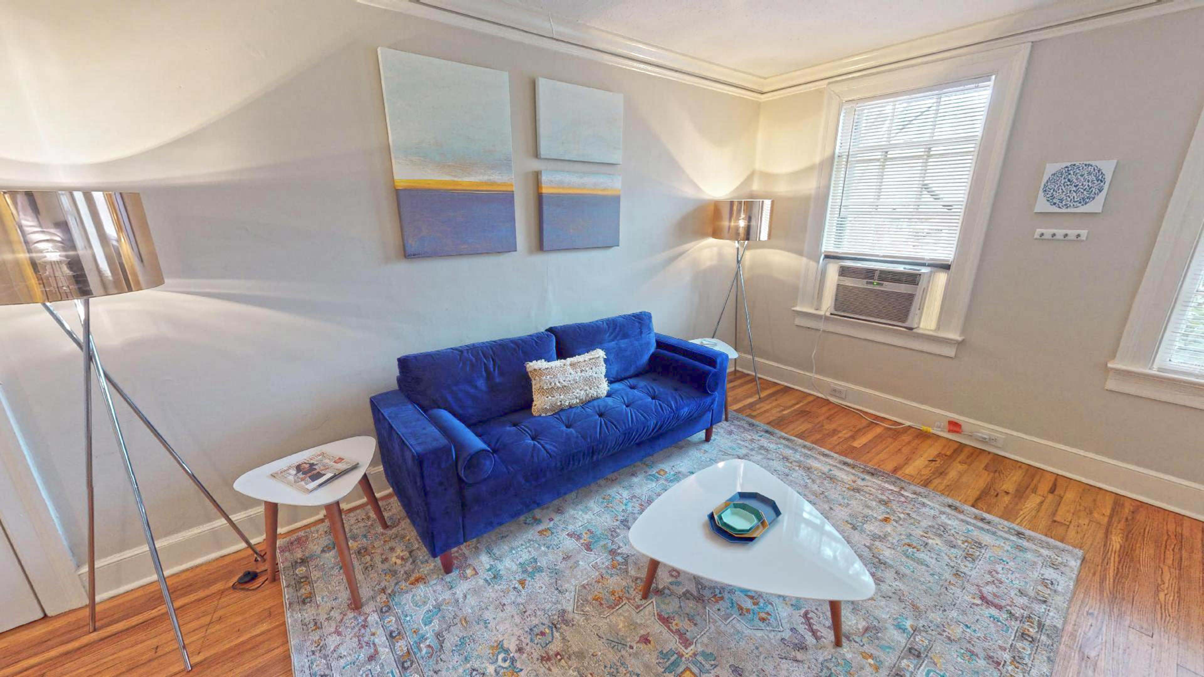 A blue velvet sofa sits beside a small white table in a room with light wood flooring and abstract wall art.