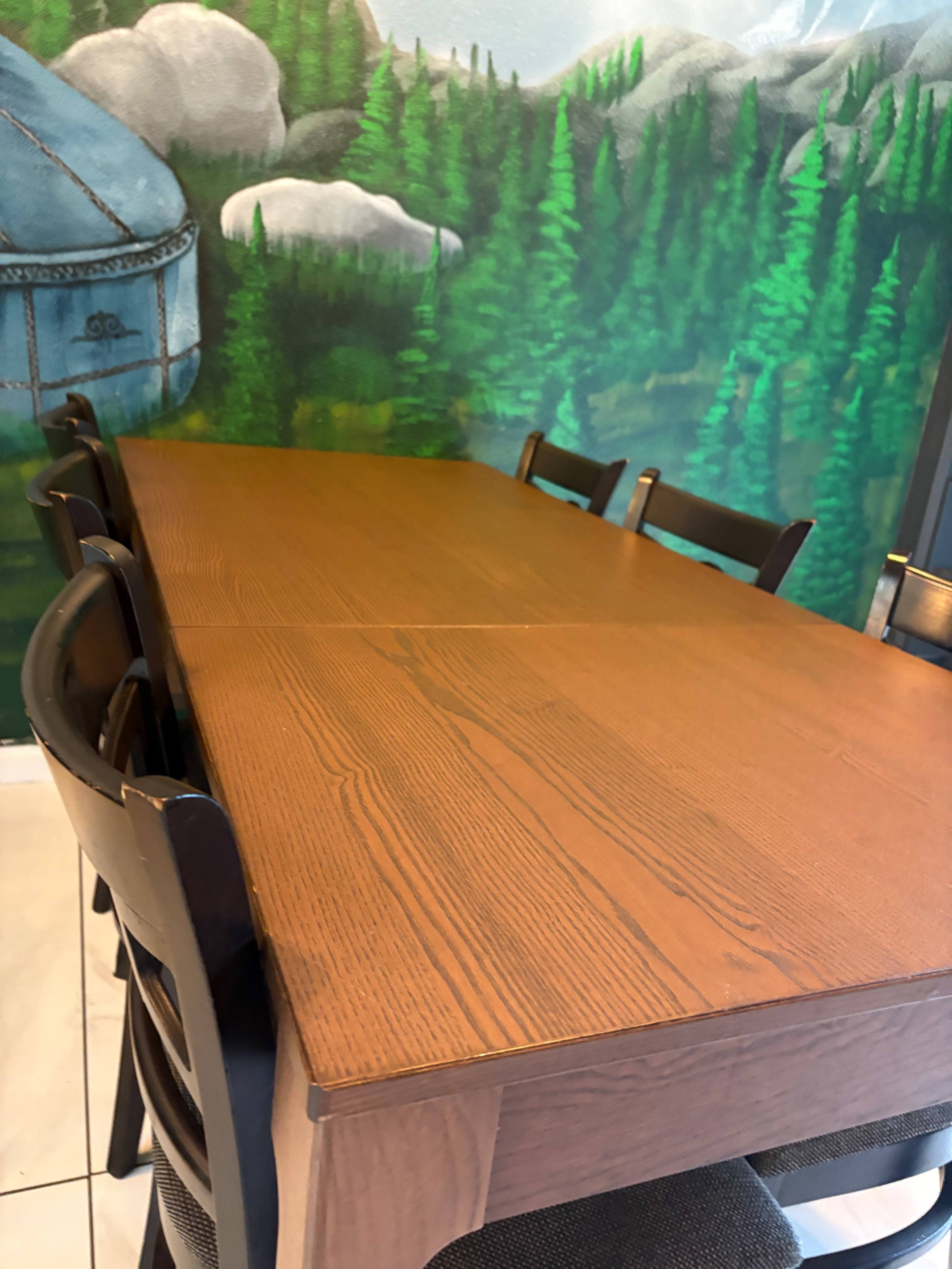 A long wooden table with black chairs is positioned in front of a mural featuring green trees and rocky mountains.