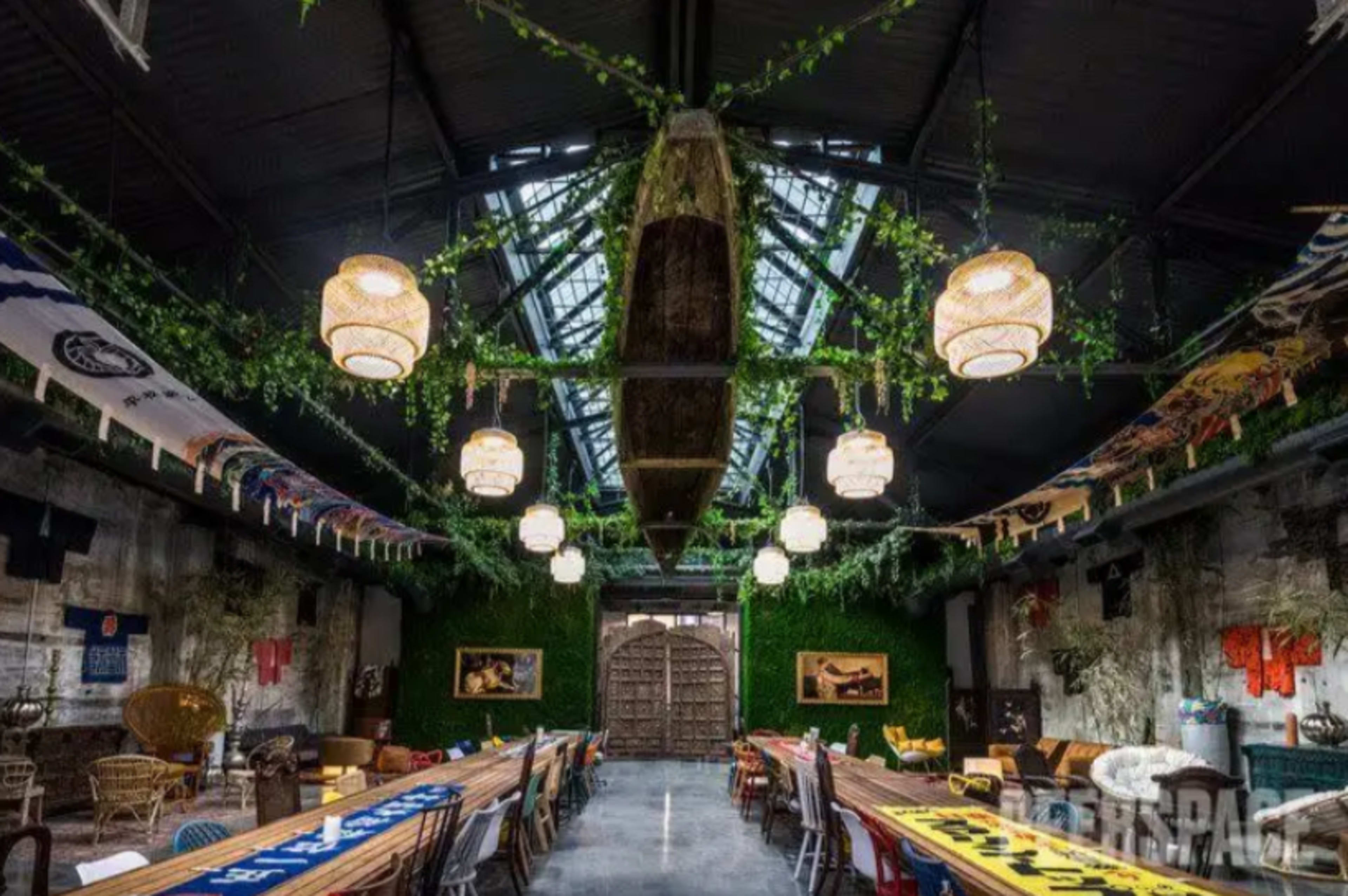 The image shows an industrial-style dining space adorned with hanging plants, lanterns, and a canoe suspended from the ceiling, featuring long communal tables arranged down the center.