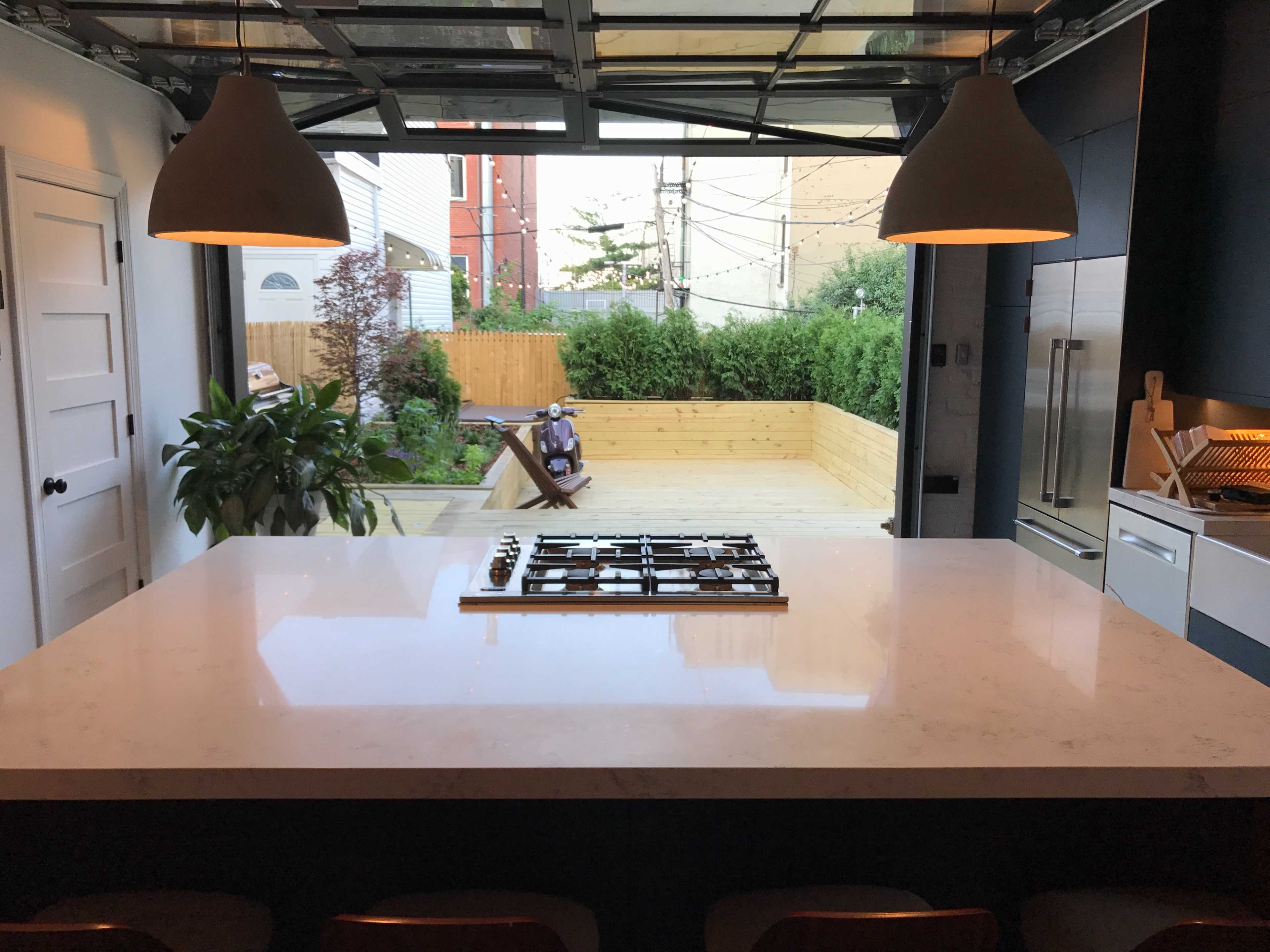 A modern kitchen features a large island with a stovetop and an open view of a backyard through a glass garage door.