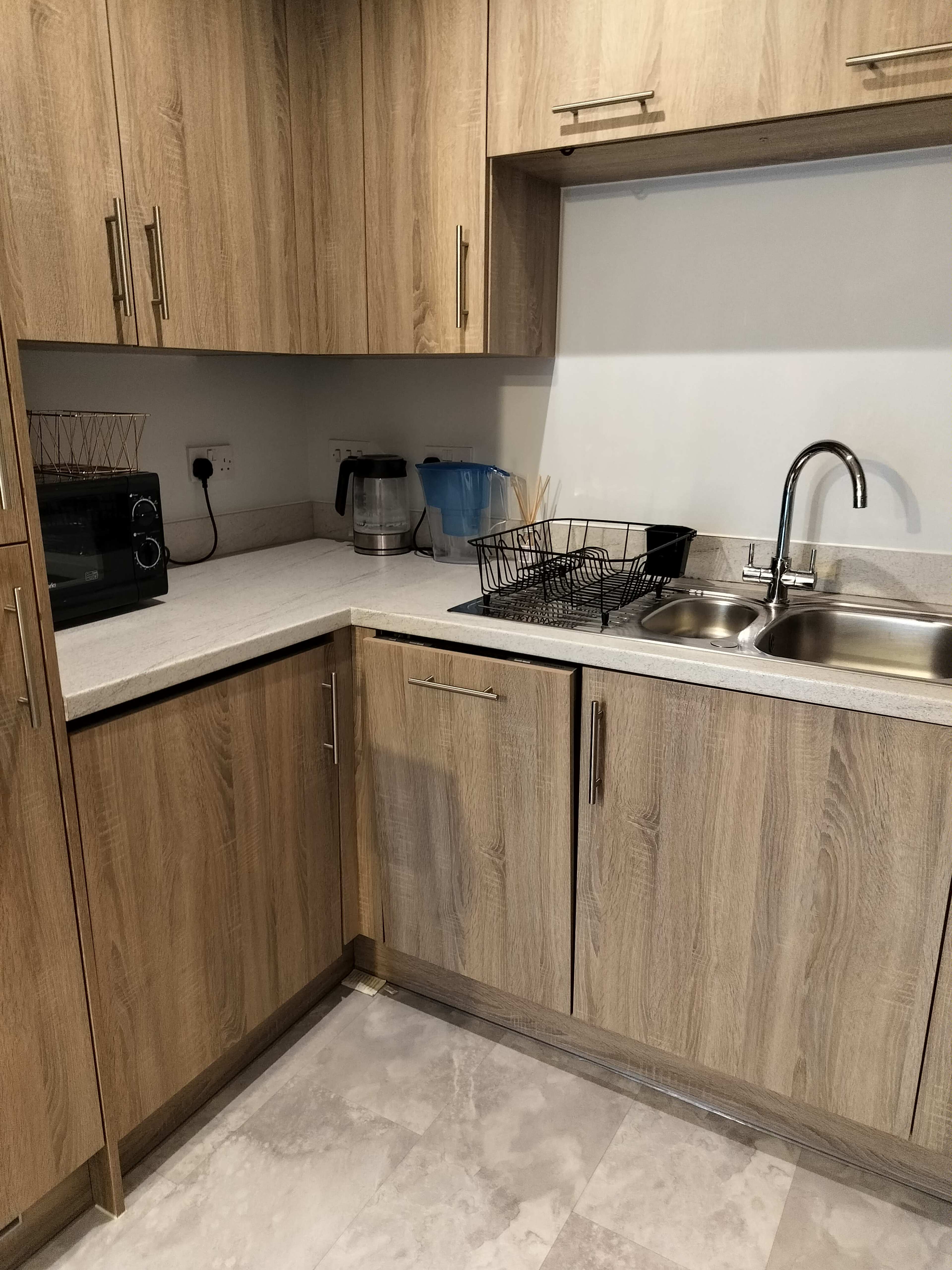The image shows a kitchen corner with wooden cabinetry, a sink, a dish drying rack, a microwave, and a water pitcher.