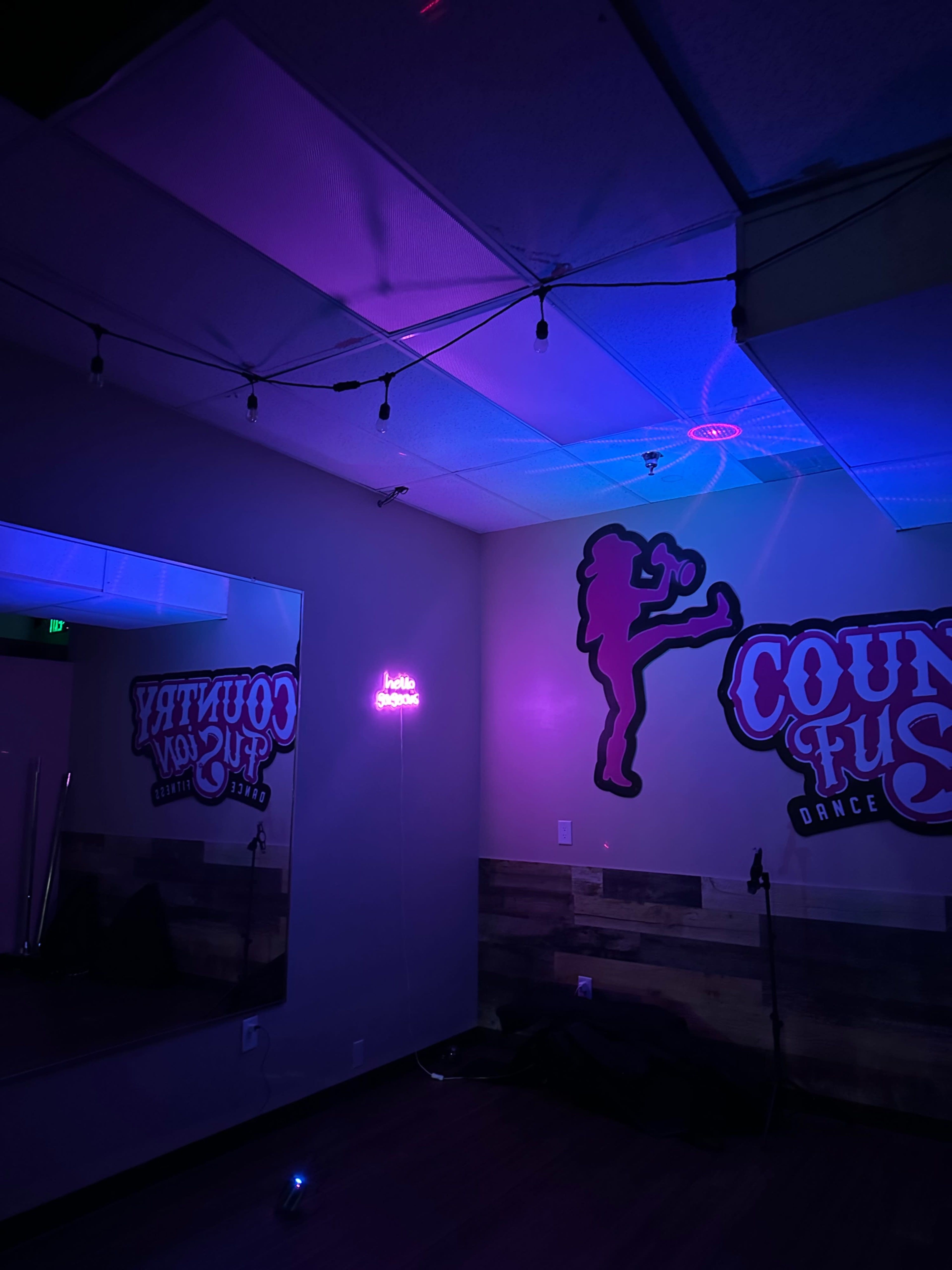 A dance studio featuring a mirrored wall, colorful LED lights casting purple and blue hues, and a neon sign that reads "hello gorgeous."
