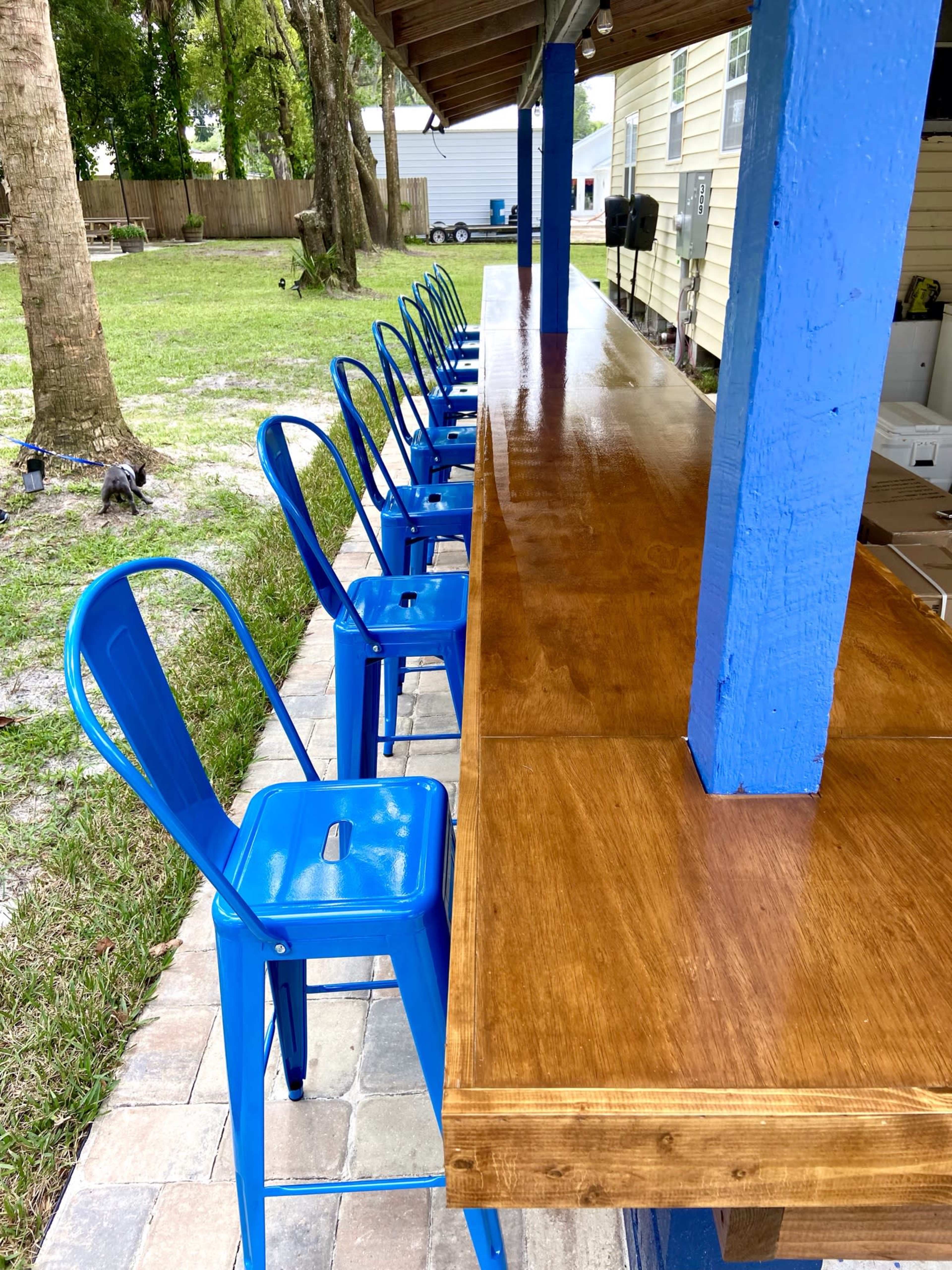 A row of blue metal chairs is positioned in front of a wooden bar under a covered outdoor area.