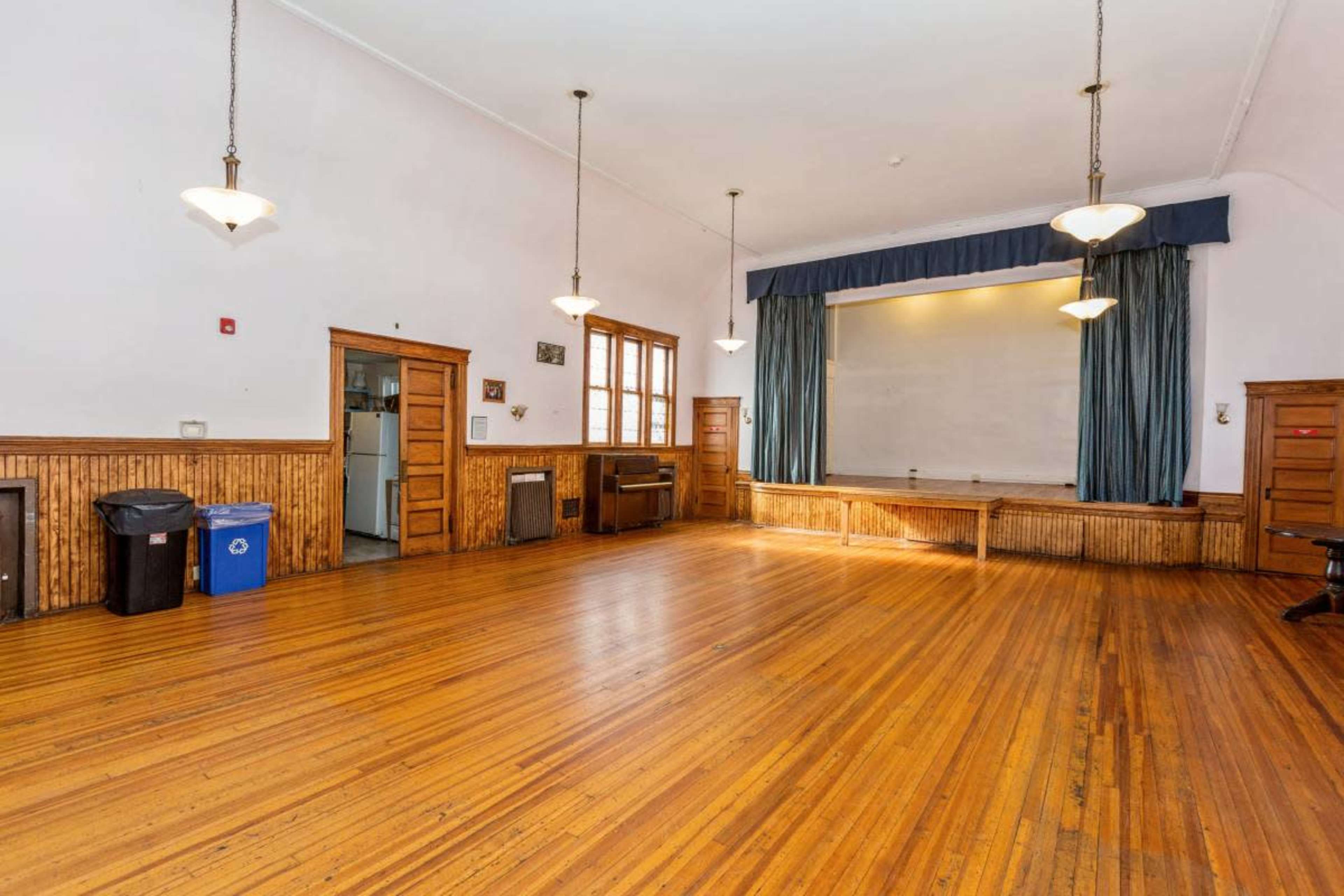 The image shows a spacious wooden-floored hall with large windows and a curtained stage at one end.