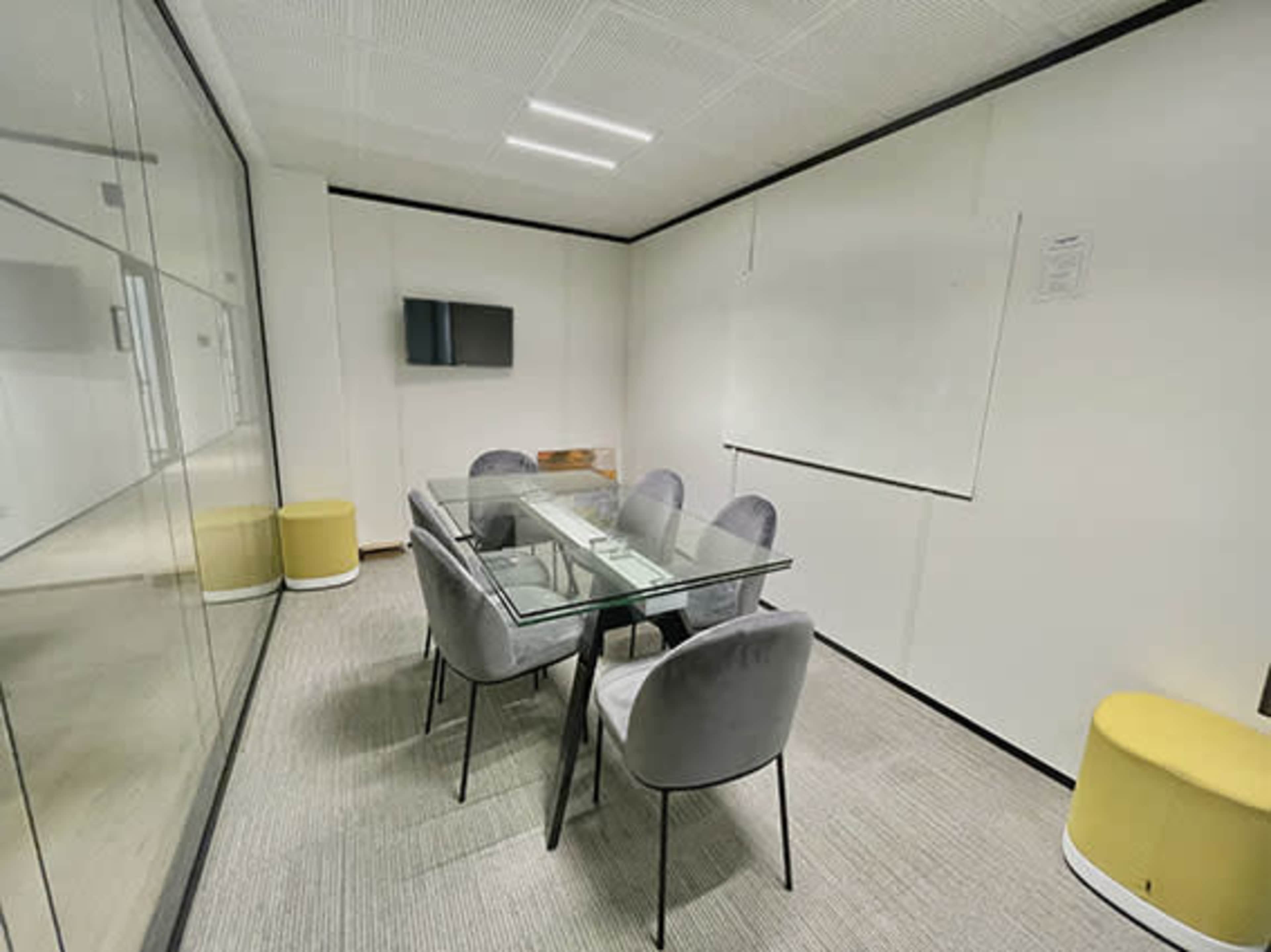A conference room features a glass table surrounded by gray chairs, with a television and a whiteboard on the walls.